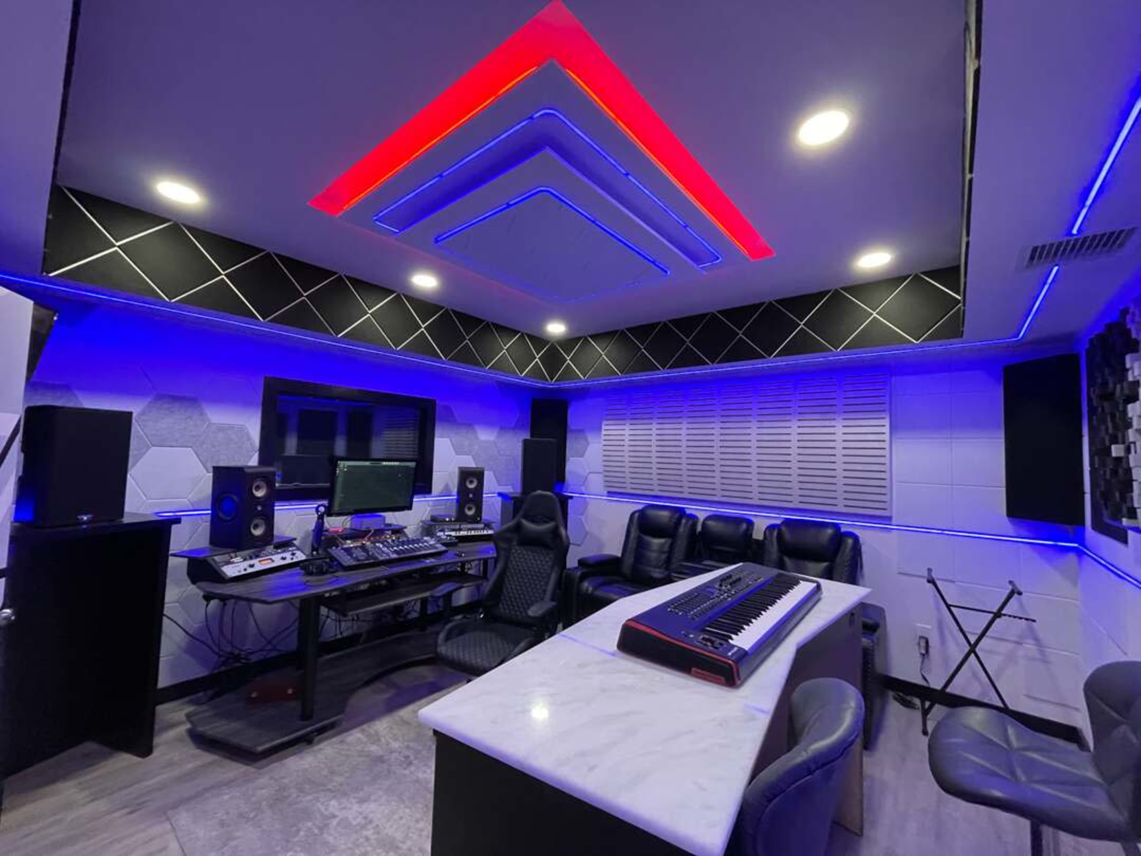 The image shows a modern music studio with a sound mixing console, various audio equipment, a keyboard, and seating arranged in a sleek design with blue and red LED lighting.