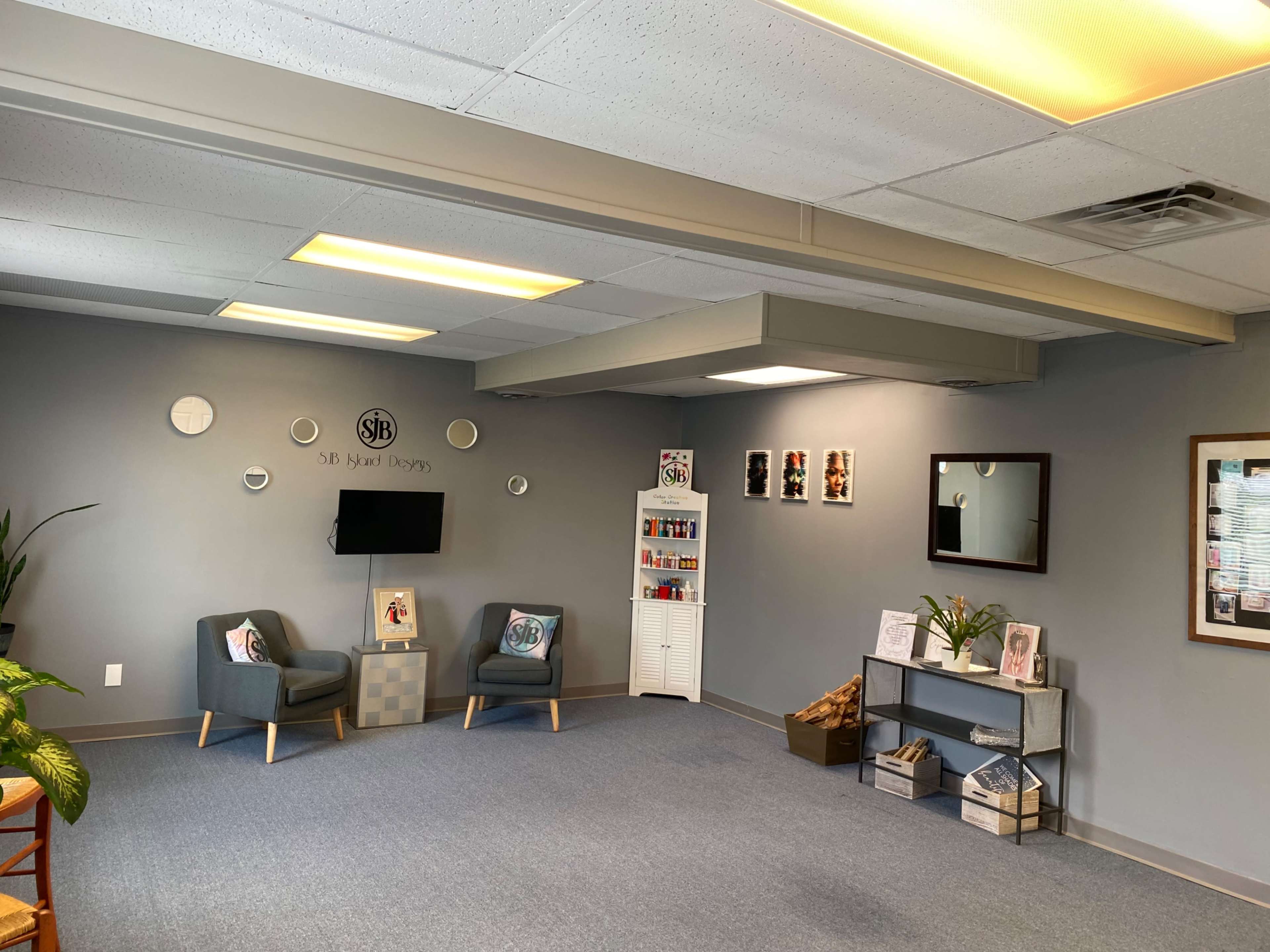 A spacious waiting area with gray walls, two armchairs, a small shelf displaying various products, a mirror, and framed artwork on the walls.