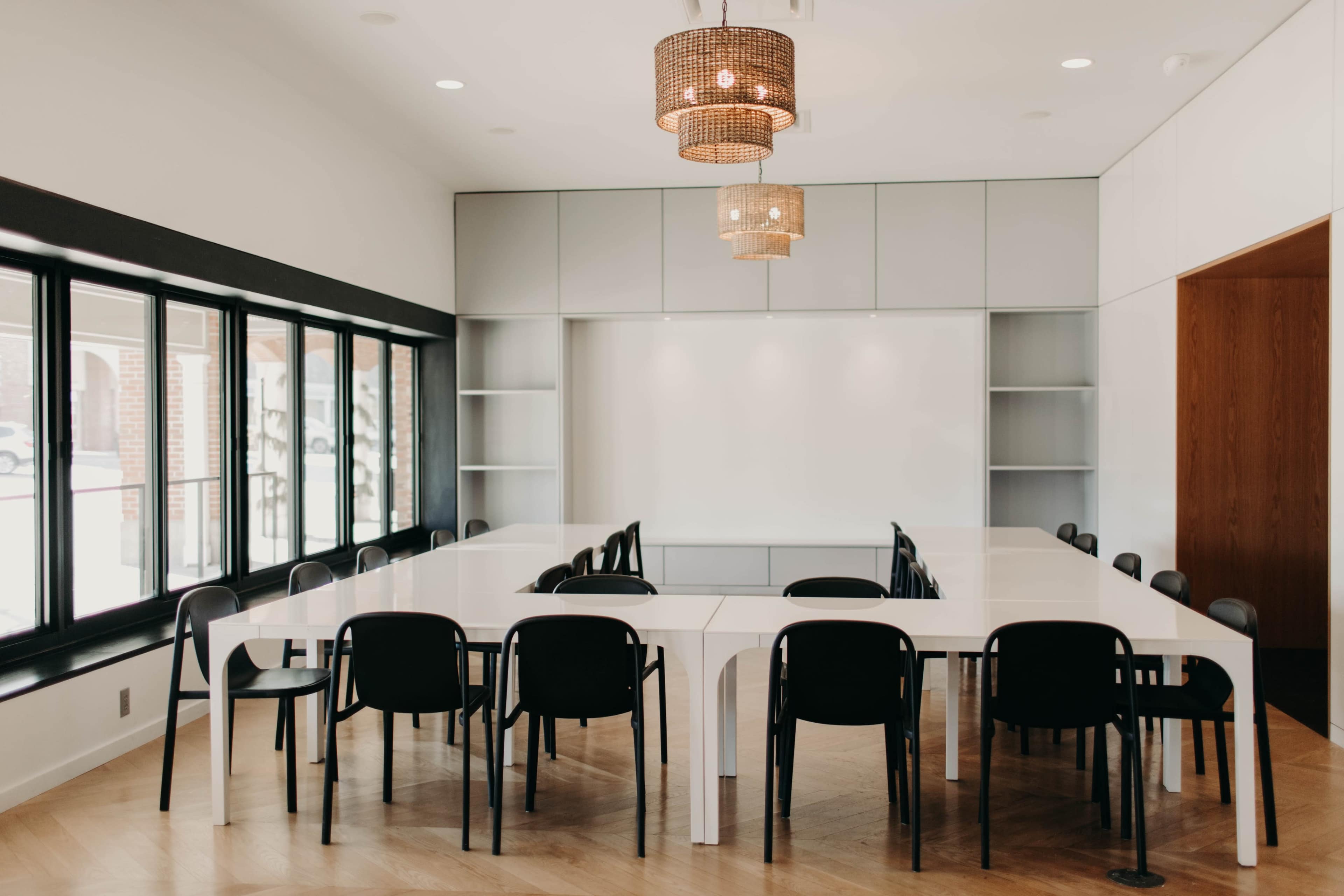 A spacious conference room features a long, white table surrounded by black chairs, with large windows and pendant lights above.