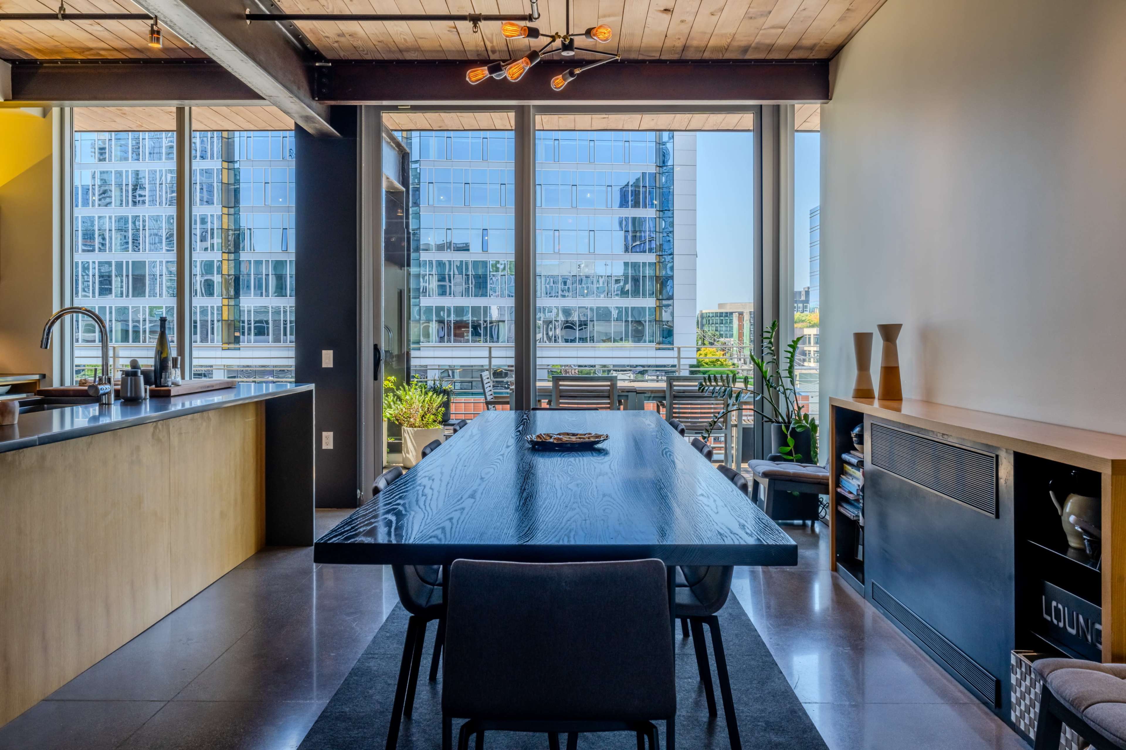 A modern dining area with a long black table surrounded by chairs, and large windows providing a view of nearby buildings and outdoor greenery.