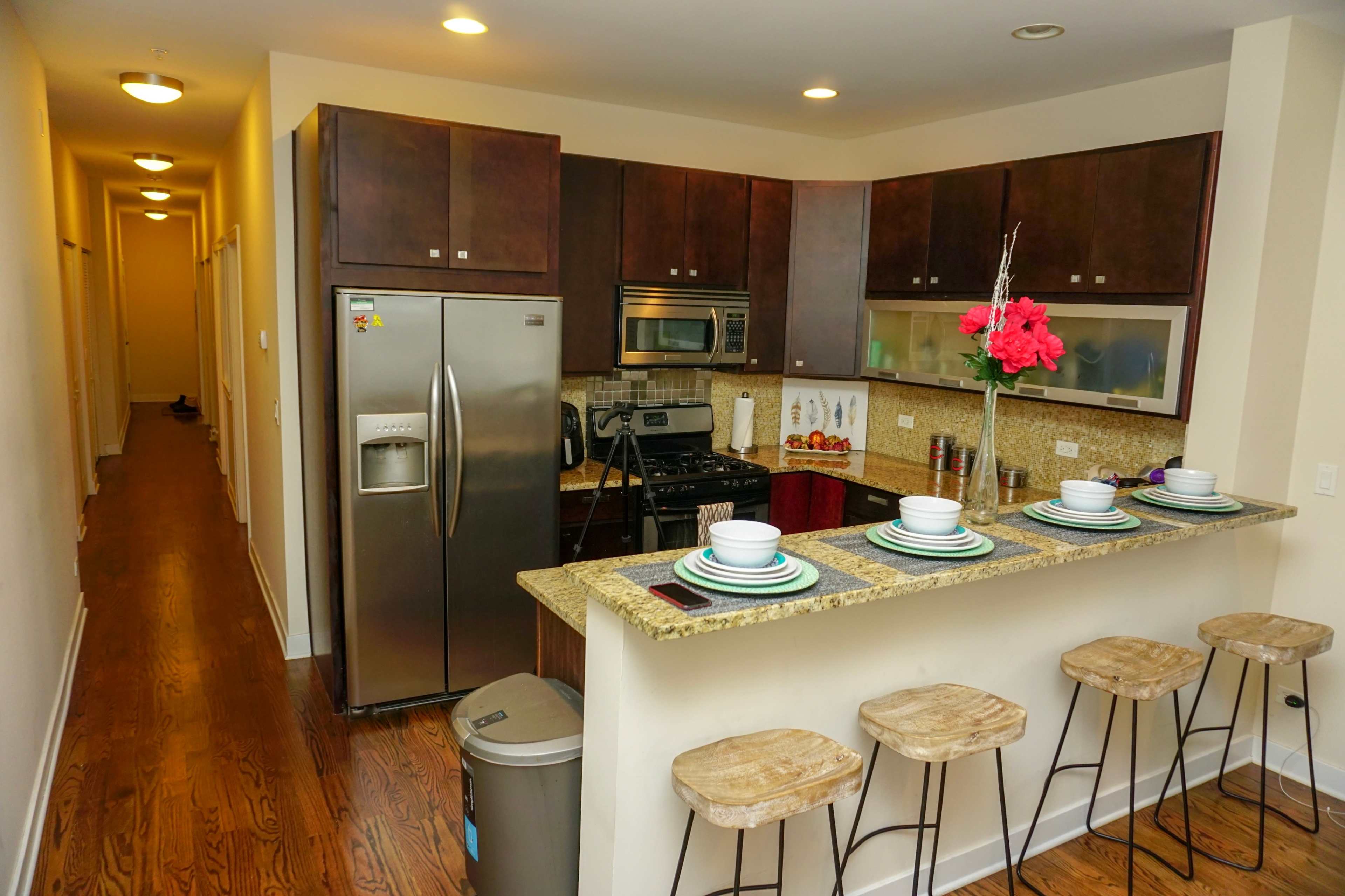 A modern kitchen with dark wooden cabinets, stainless steel appliances, and a bar counter with four stools, overlooking a narrow hallway.