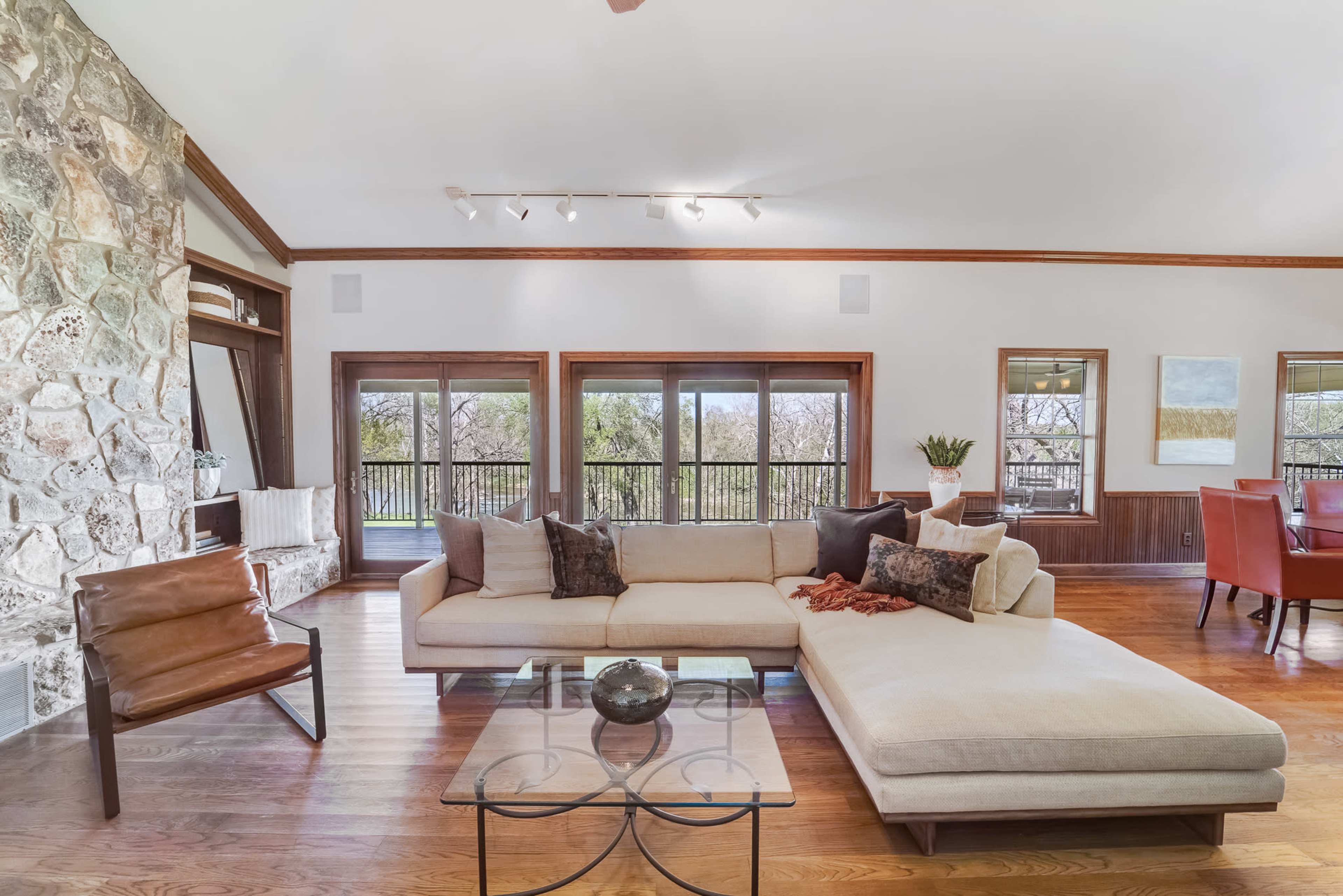 A spacious living room features a large sectional sofa, a glass coffee table, and a stone fireplace, with large windows offering views of the outdoors.