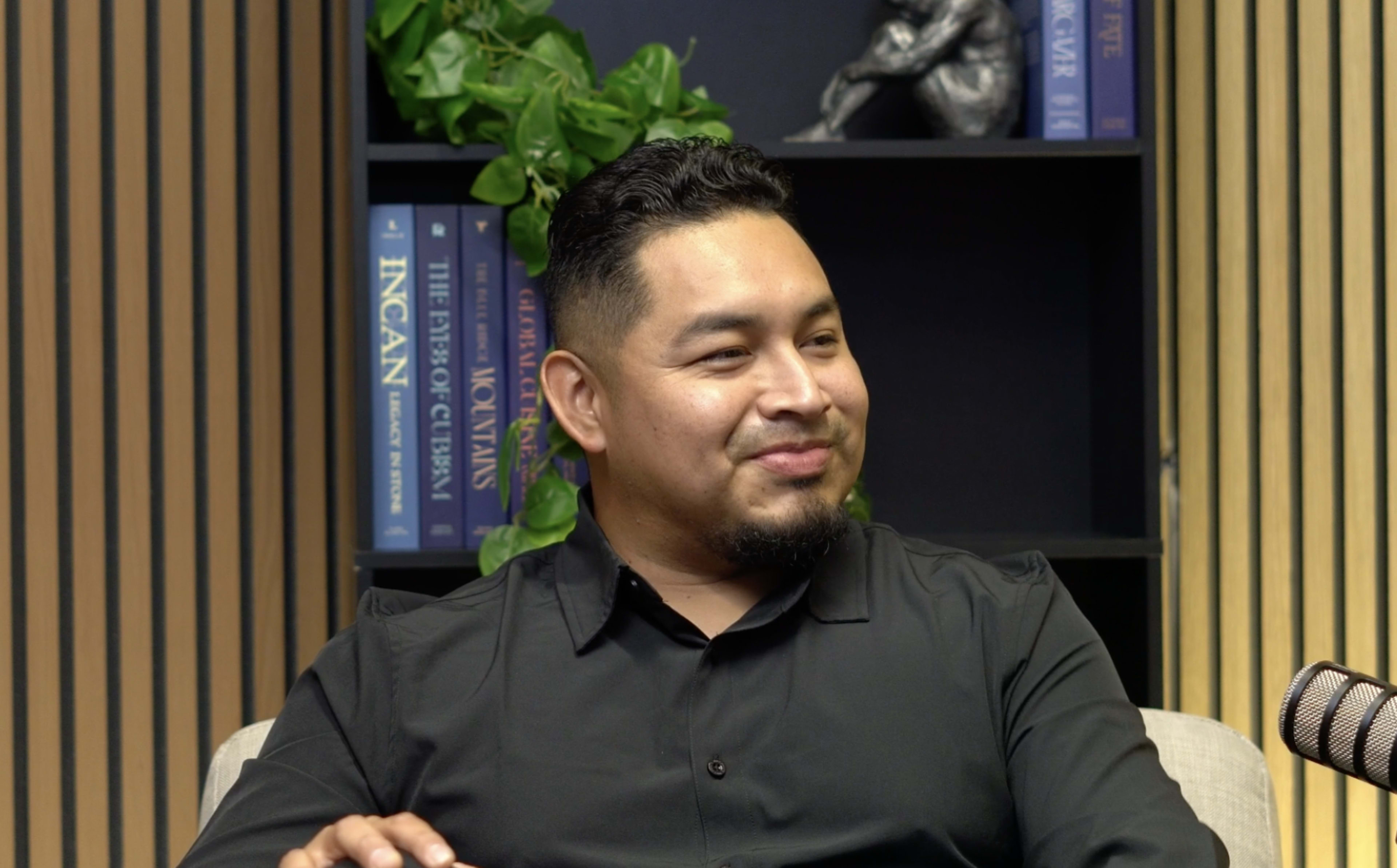 A man in a black shirt is seated at a table, smiling while facing the camera, with a bookshelf filled with various books in the background.