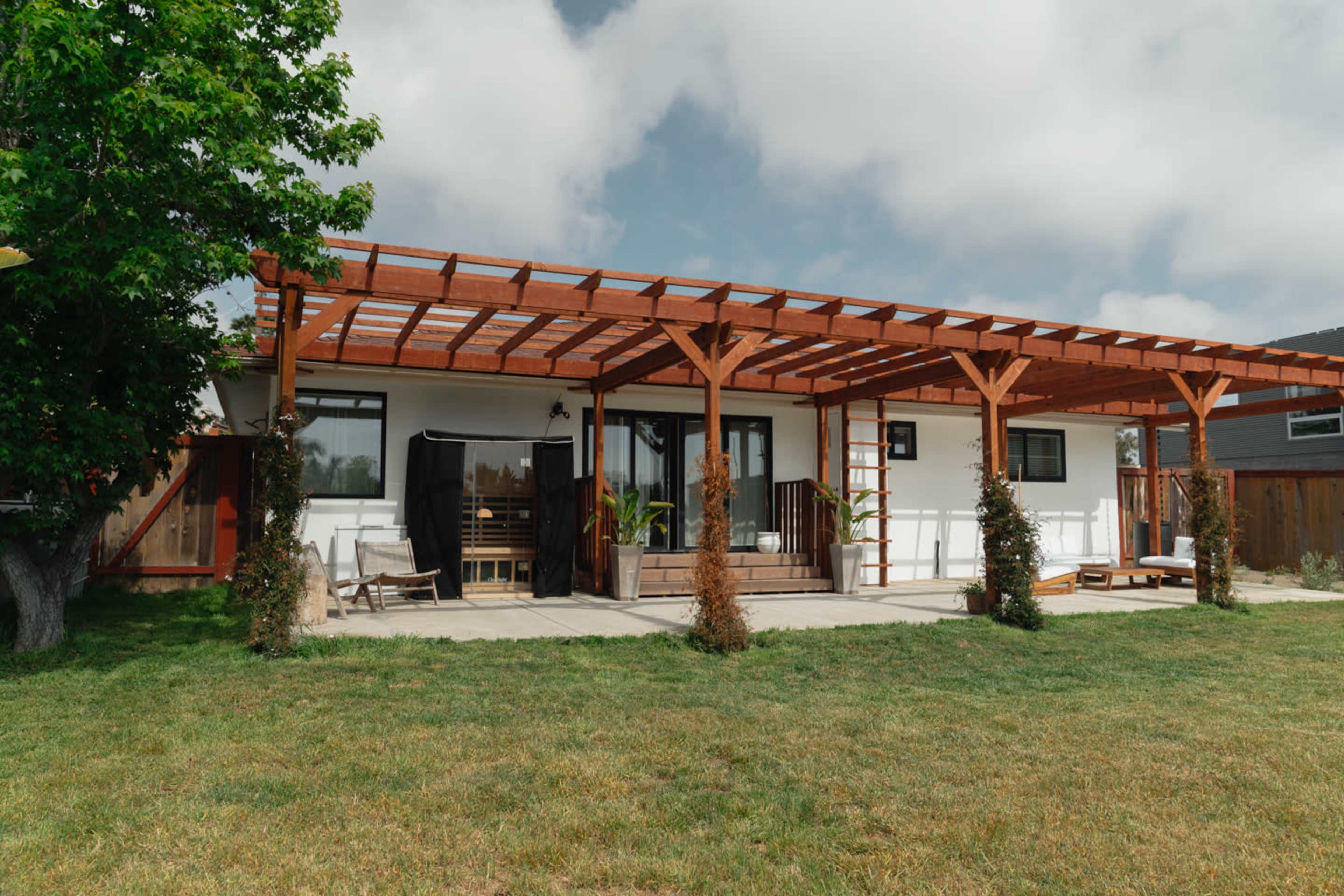 A modern house with a wooden pergola and outdoor seating area is set against a grassy lawn.