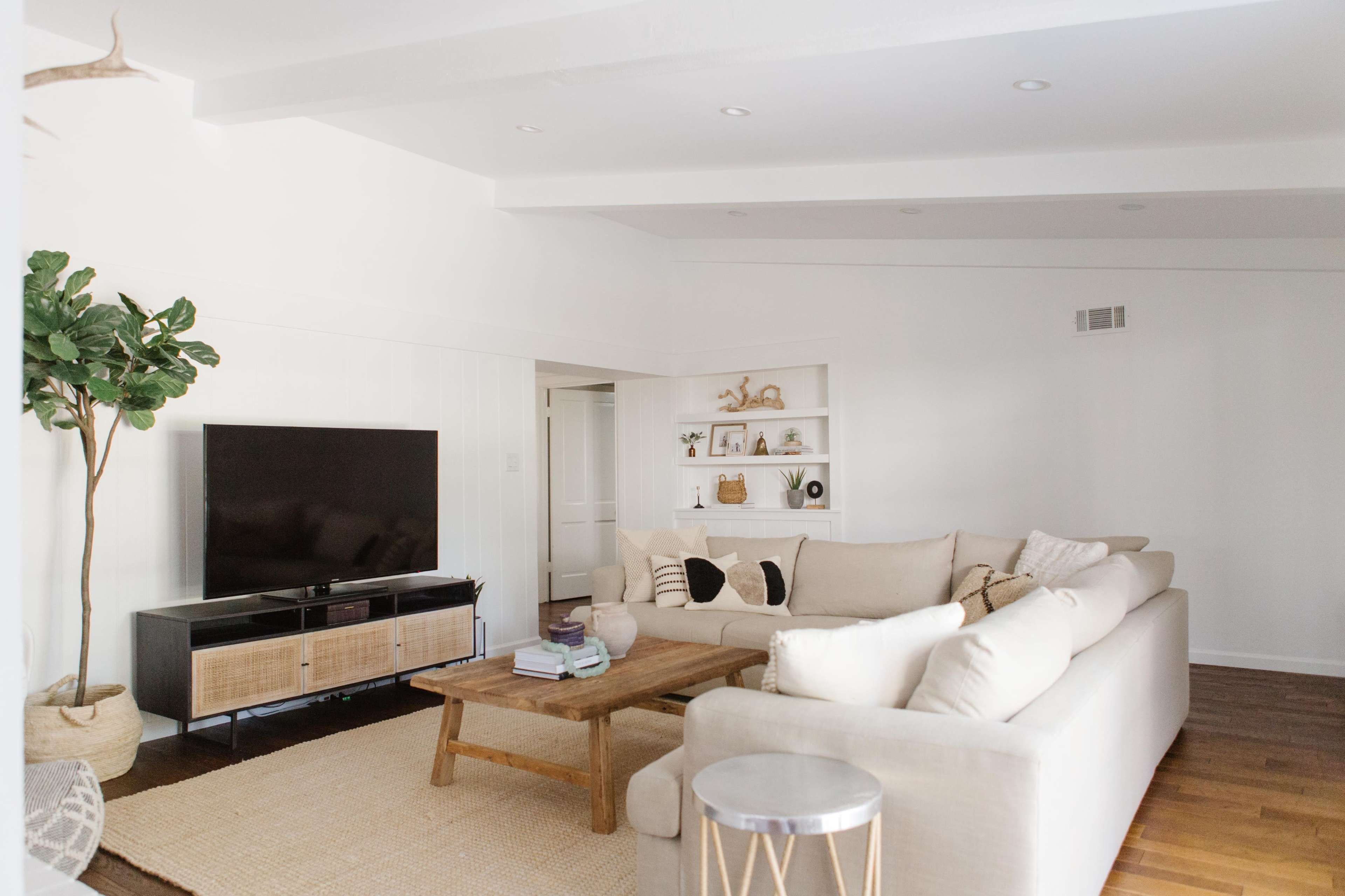 A spacious living room features a large sectional sofa, a coffee table, a television on a stand, and a potted plant, all arranged around a light-colored area rug.