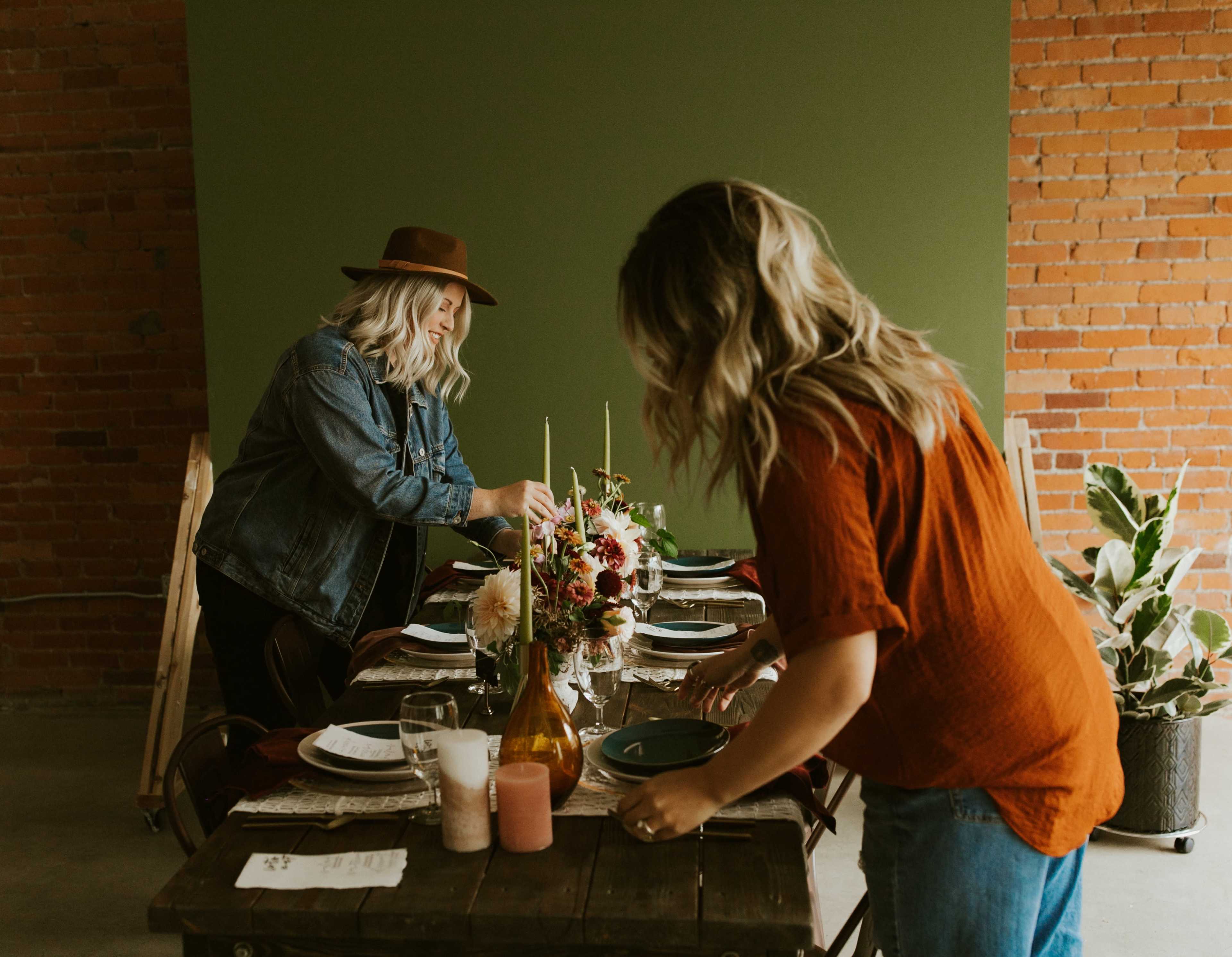 Two women are arranging table settings and flower centerpieces on a wooden dining table in a rustic interior space.