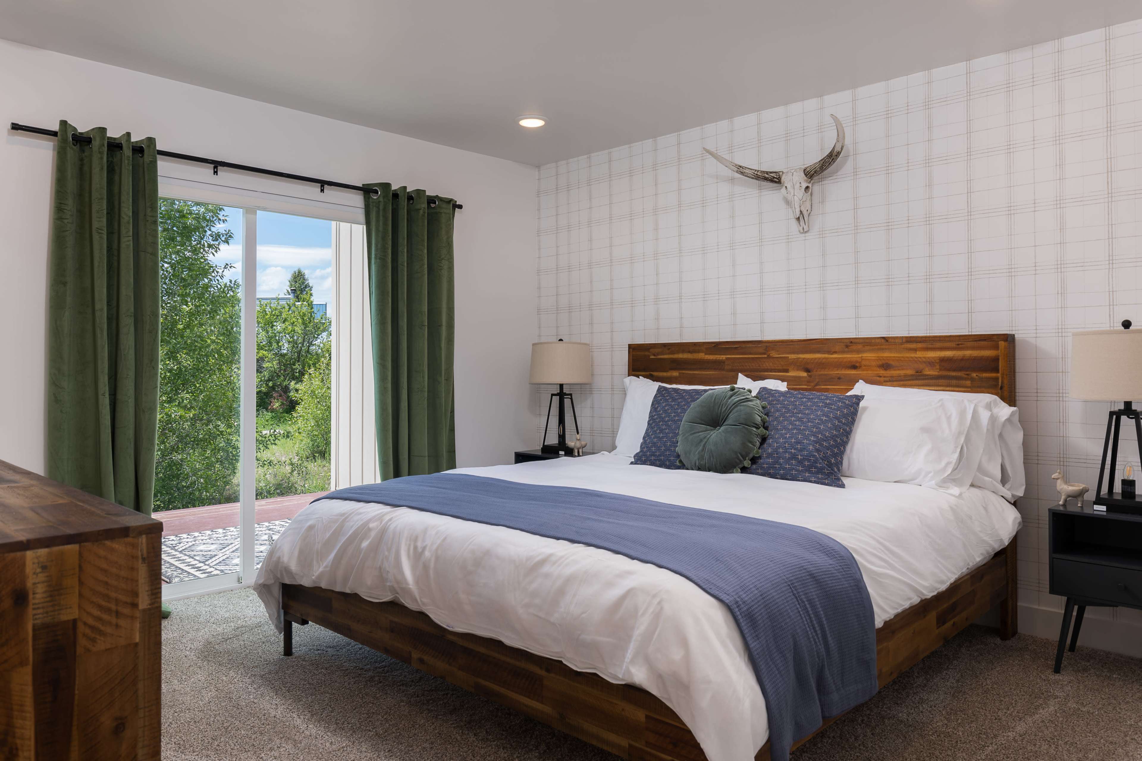 A cozy bedroom with a wooden bed, white bedding, and green curtains, featuring a patterned wall and a view through sliding glass doors.