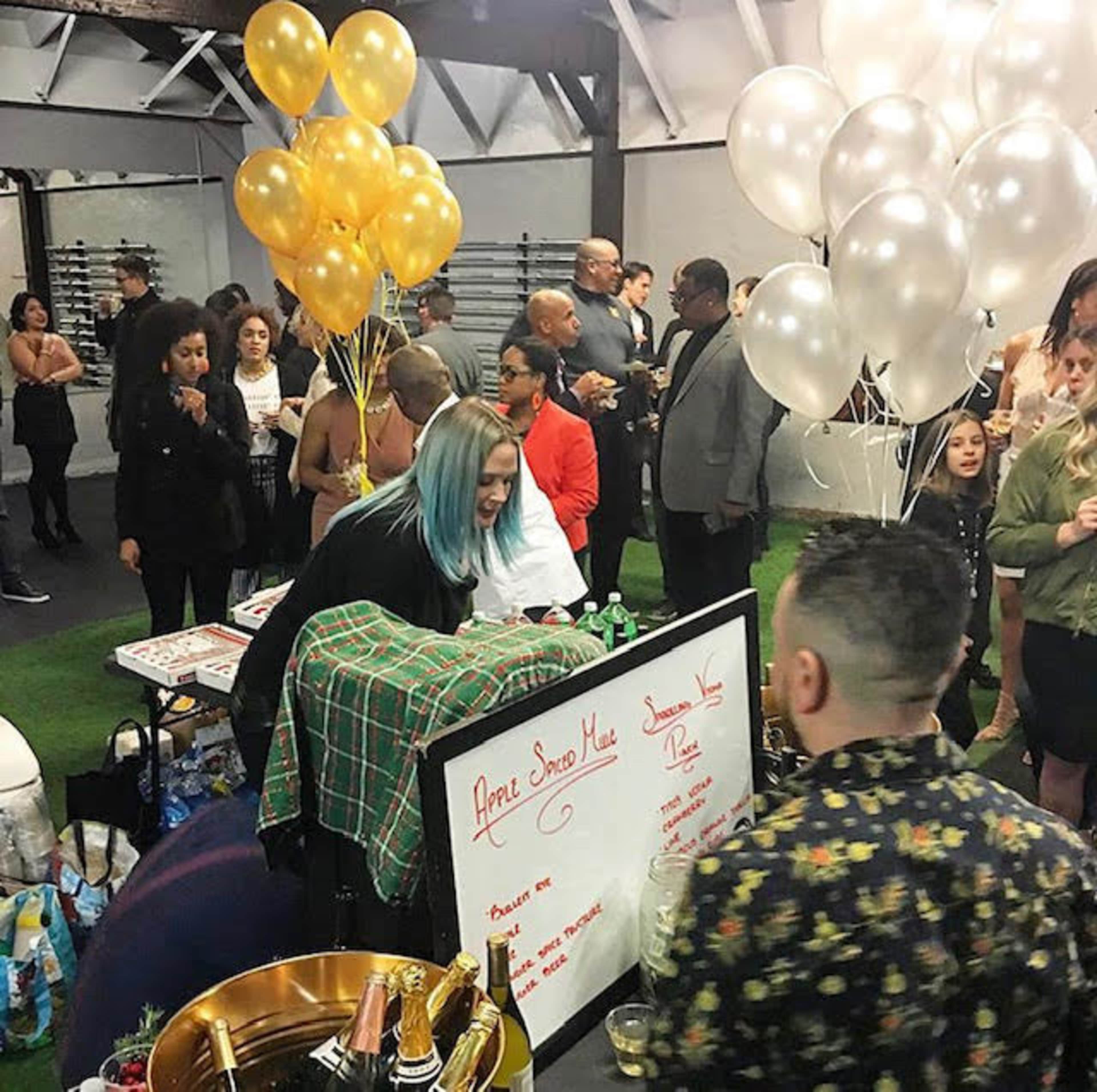A group of people socialize at a festive indoor event, with a table displaying a menu and decorative balloons in the background.