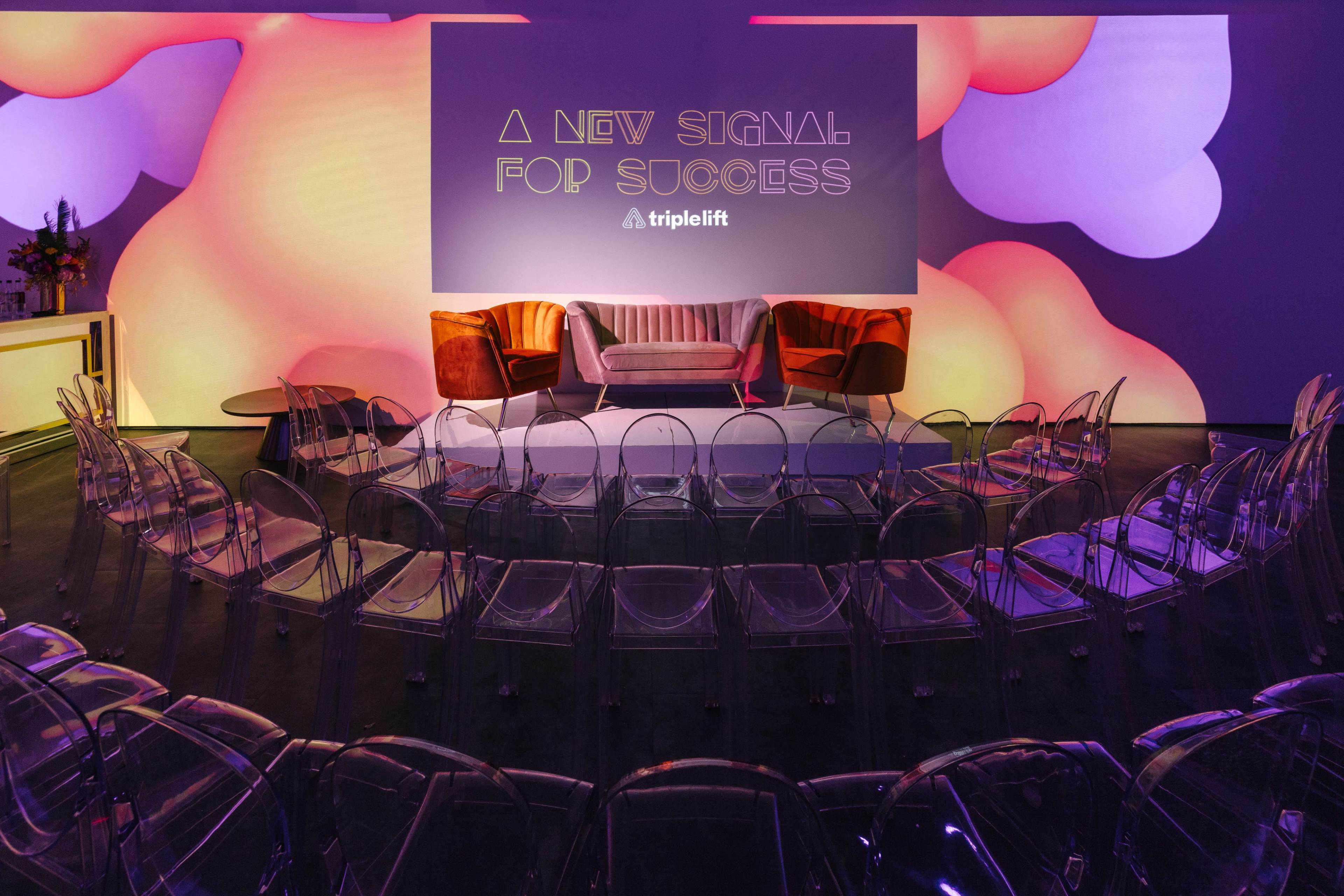 A modern event space features a circular arrangement of clear chairs facing a central stage with a backdrop that reads "A NEW SIGNAL FOR SUCCESS" along with the triplelift logo.