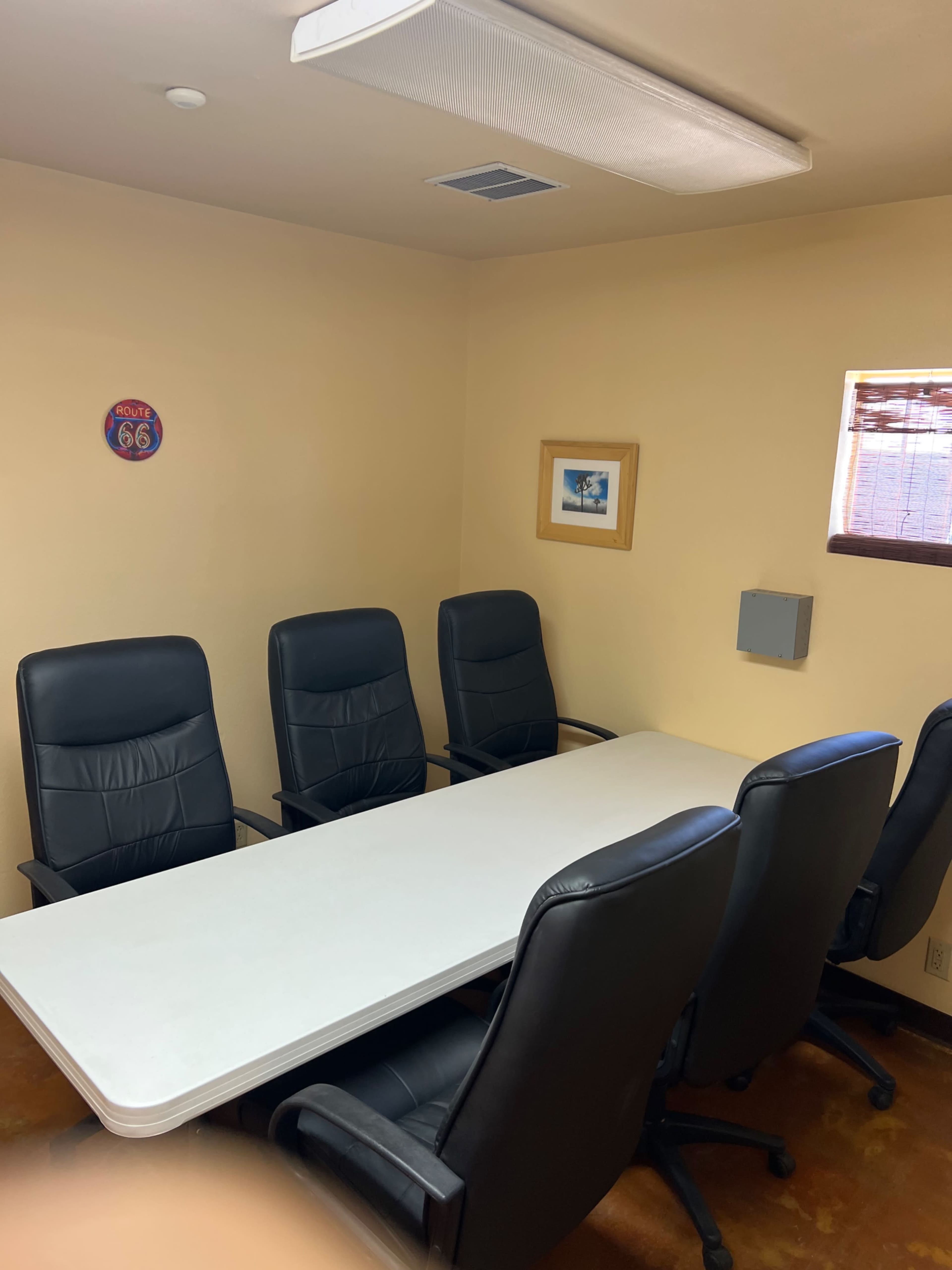 The image shows a small conference room with a long white table and four black chairs arranged around it, along with a wall decoration and a framed picture.