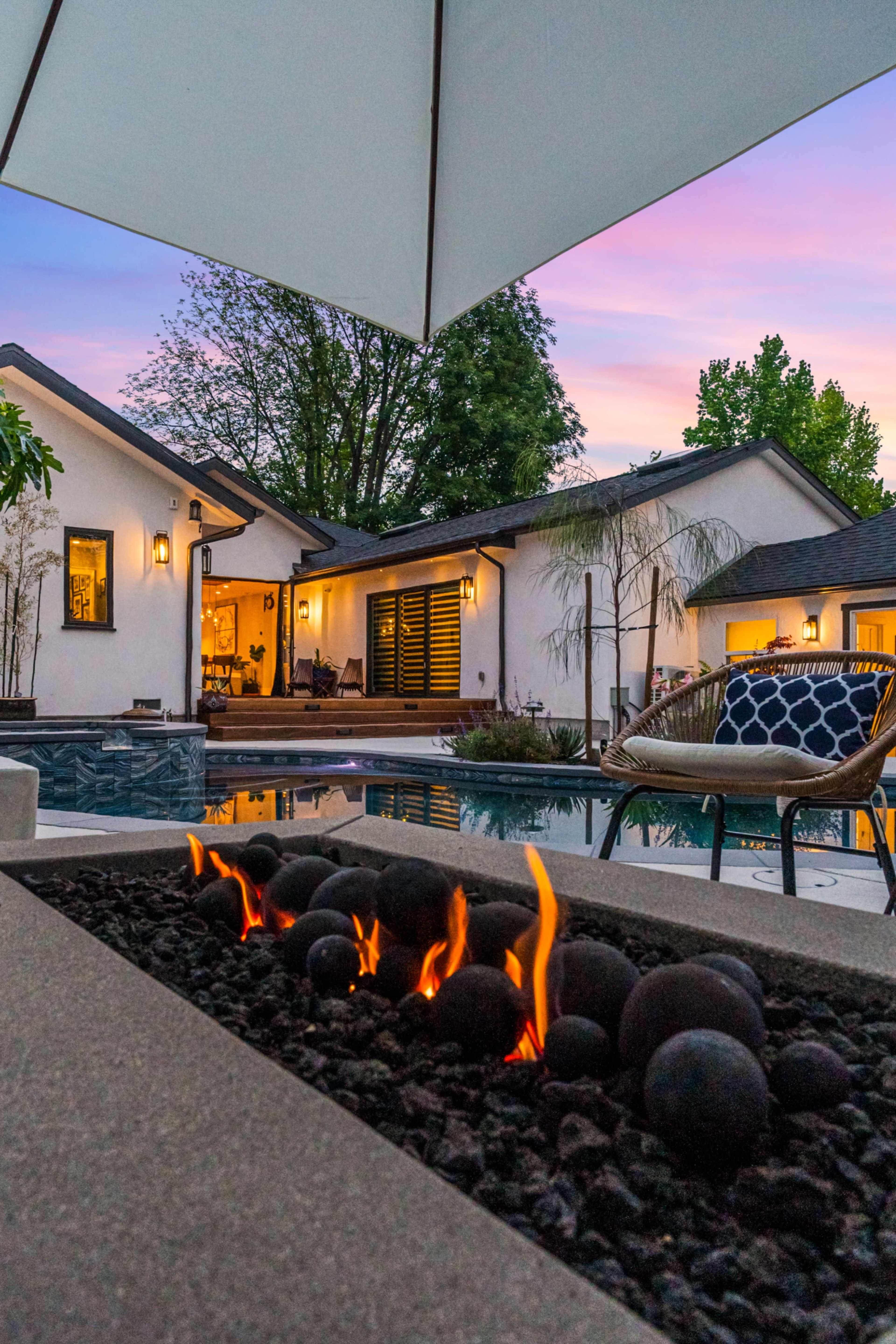 A modern backyard features a fire pit with glowing embers, a swimming pool, and a contemporary house in the background during sunset.