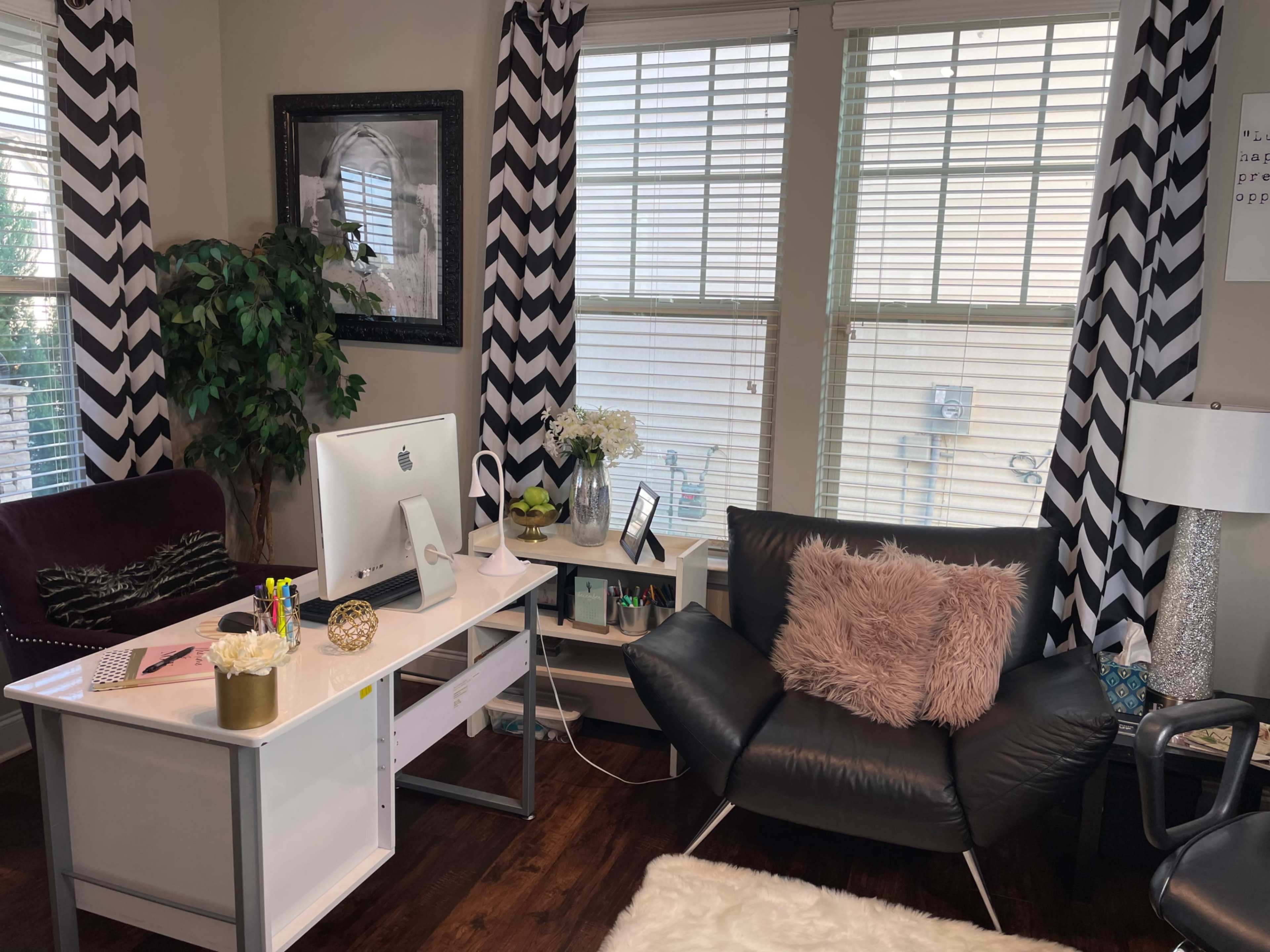 A modern office space features a white desk with an iMac, a black armchair with a pink pillow, and zigzag patterned curtains.
