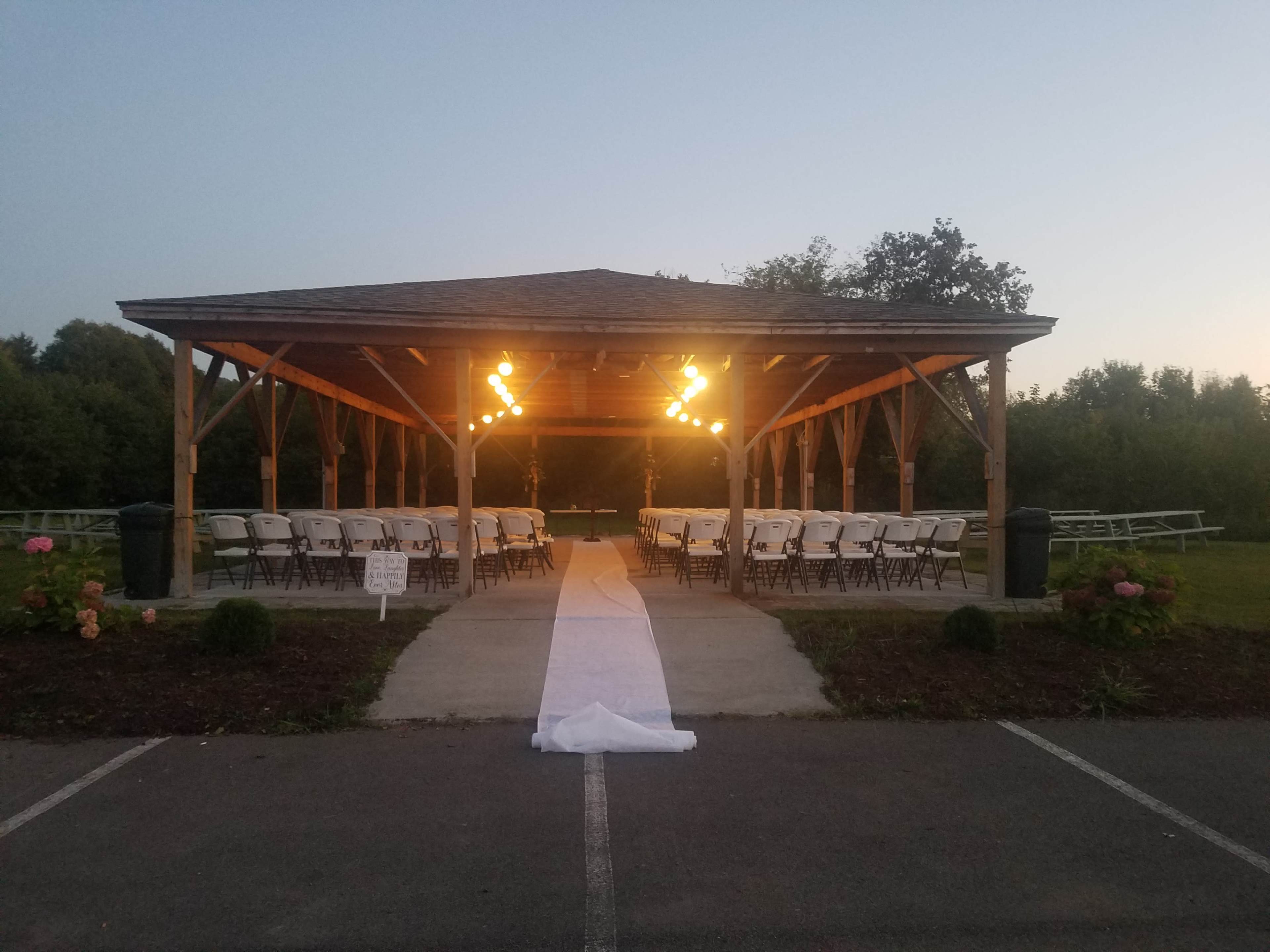 A covered pavilion is set up with rows of white chairs and a white carpet leading to the center, illuminated by overhead lights, surrounded by greenery.