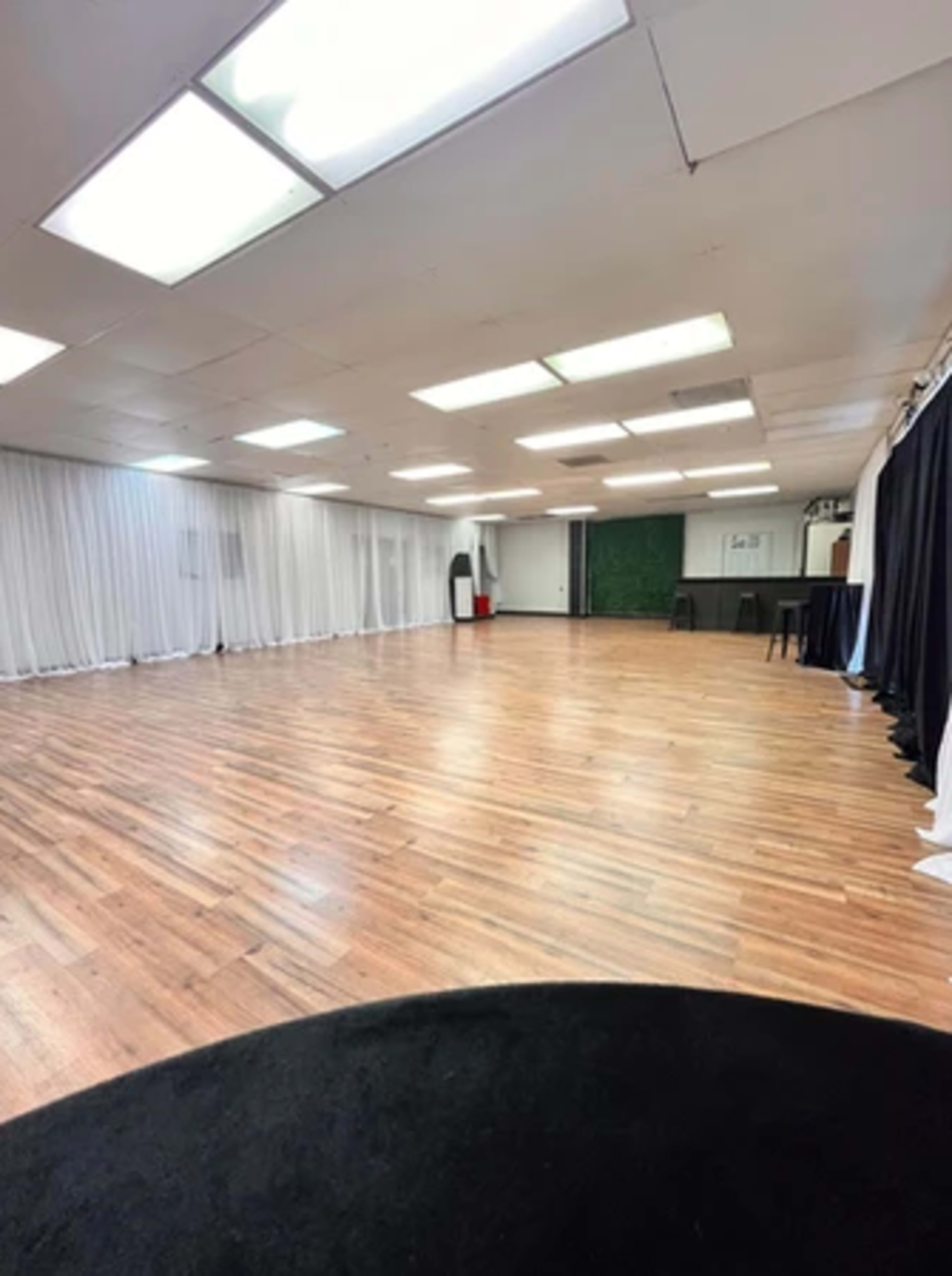 A spacious, well-lit room with wooden floors, white curtains, and a black backdrop, equipped for events or activities.