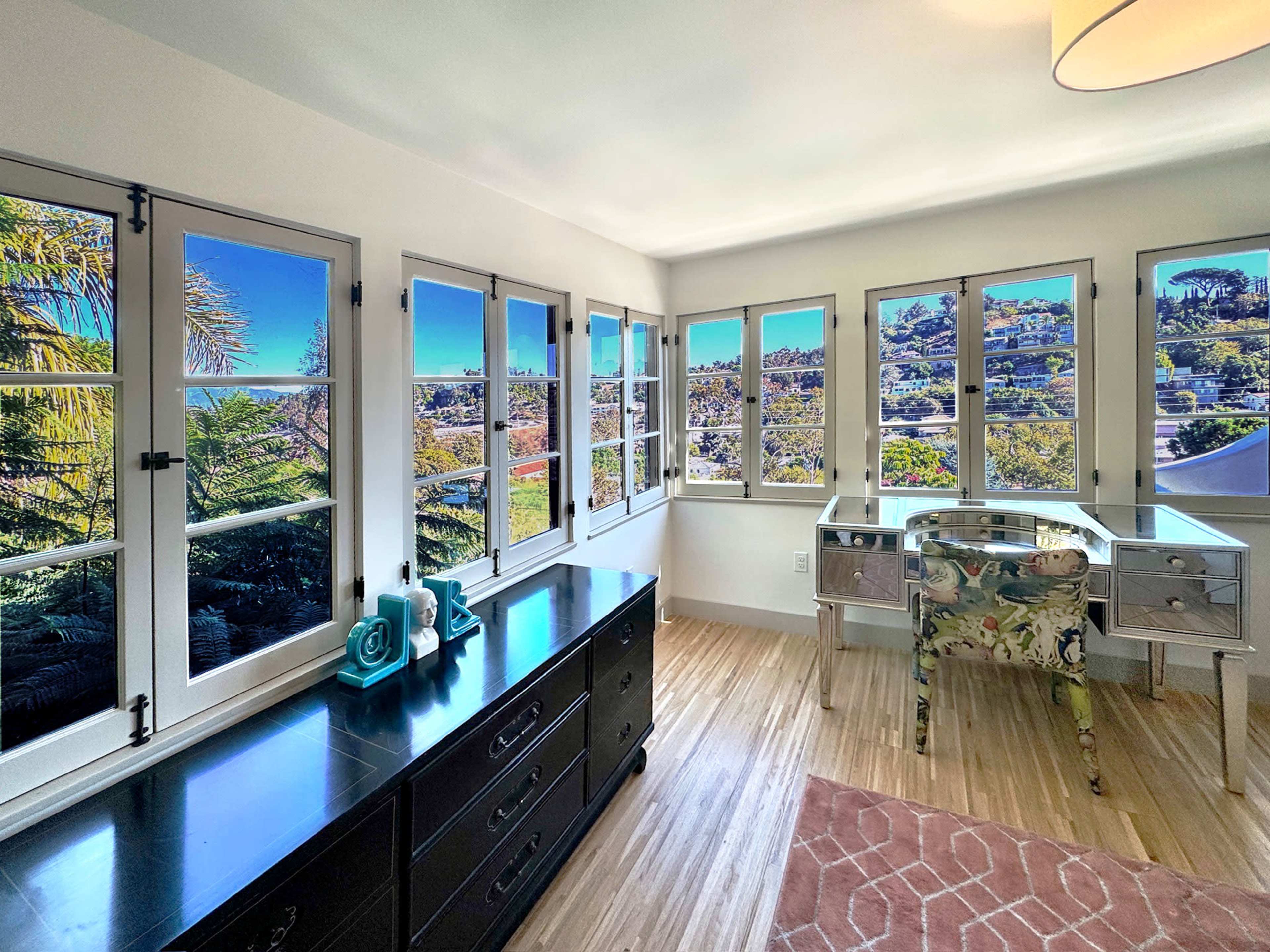 A bright room features a modern desk and a dark dresser, with large windows offering a panoramic view of the surrounding landscape.