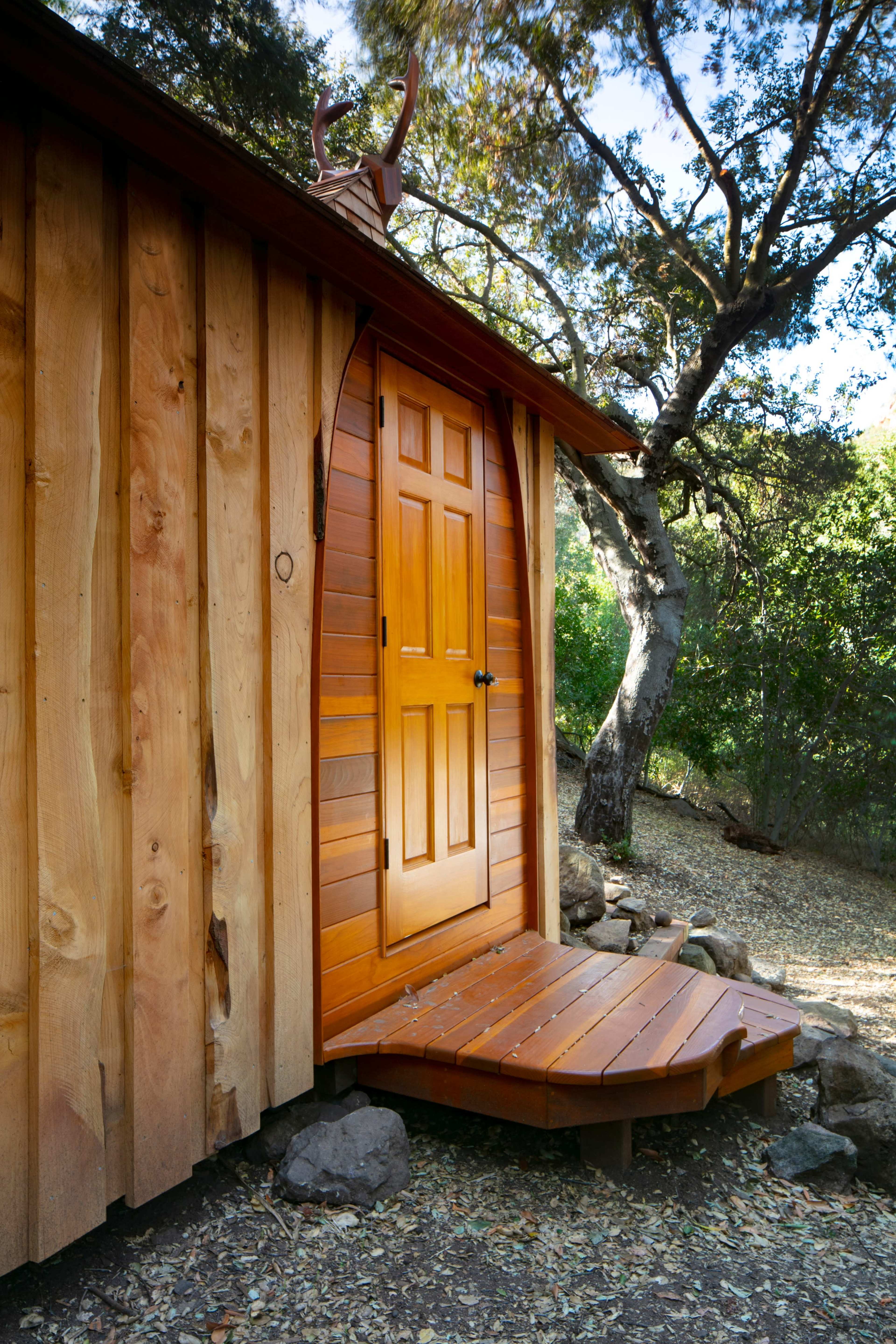 A wooden cabin with a curved door and a small deck sits among trees in a wooded area.