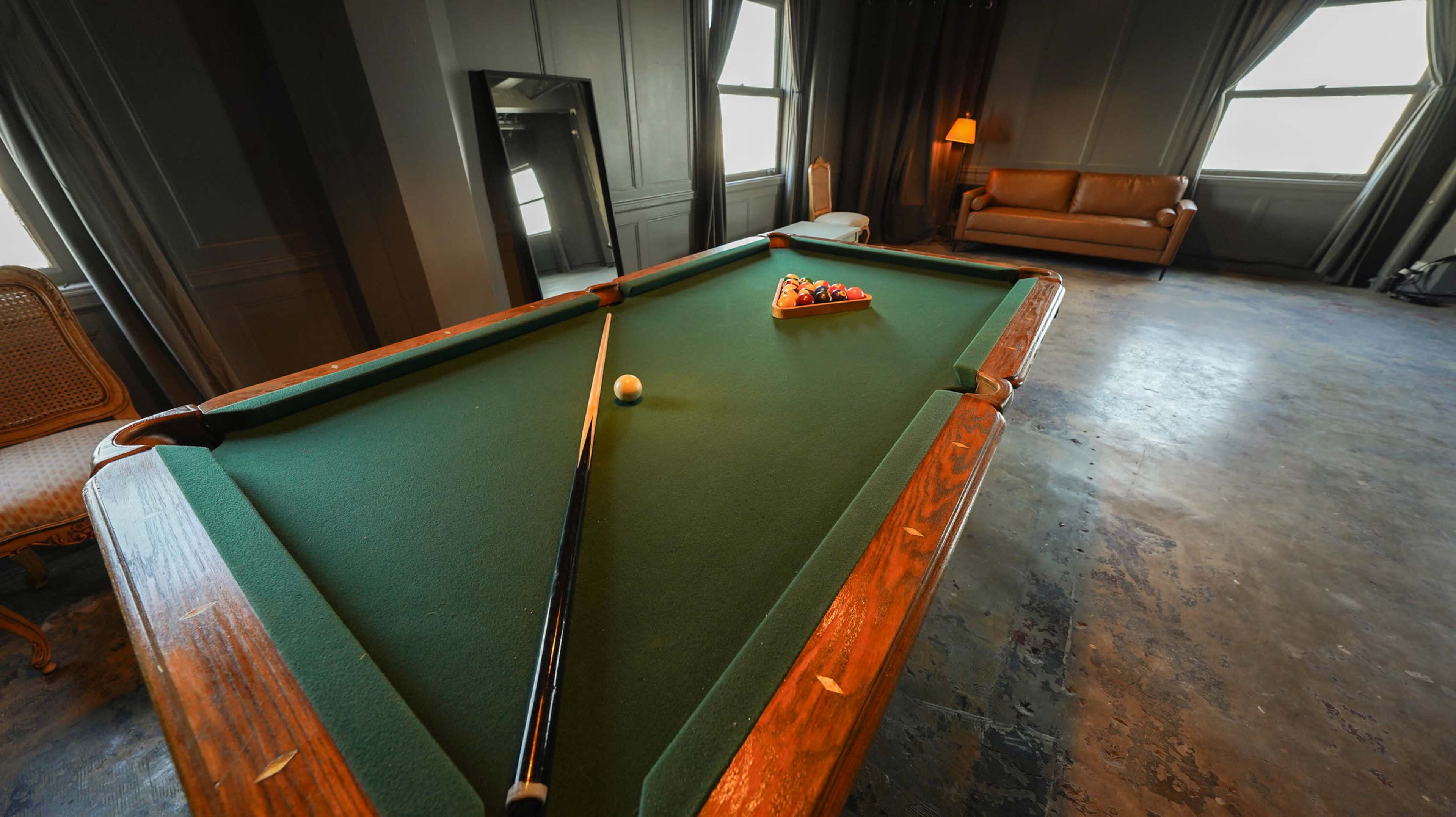 A billiards table with a cue stick and a cue ball is centered in a dimly lit room featuring large windows and a couch.
