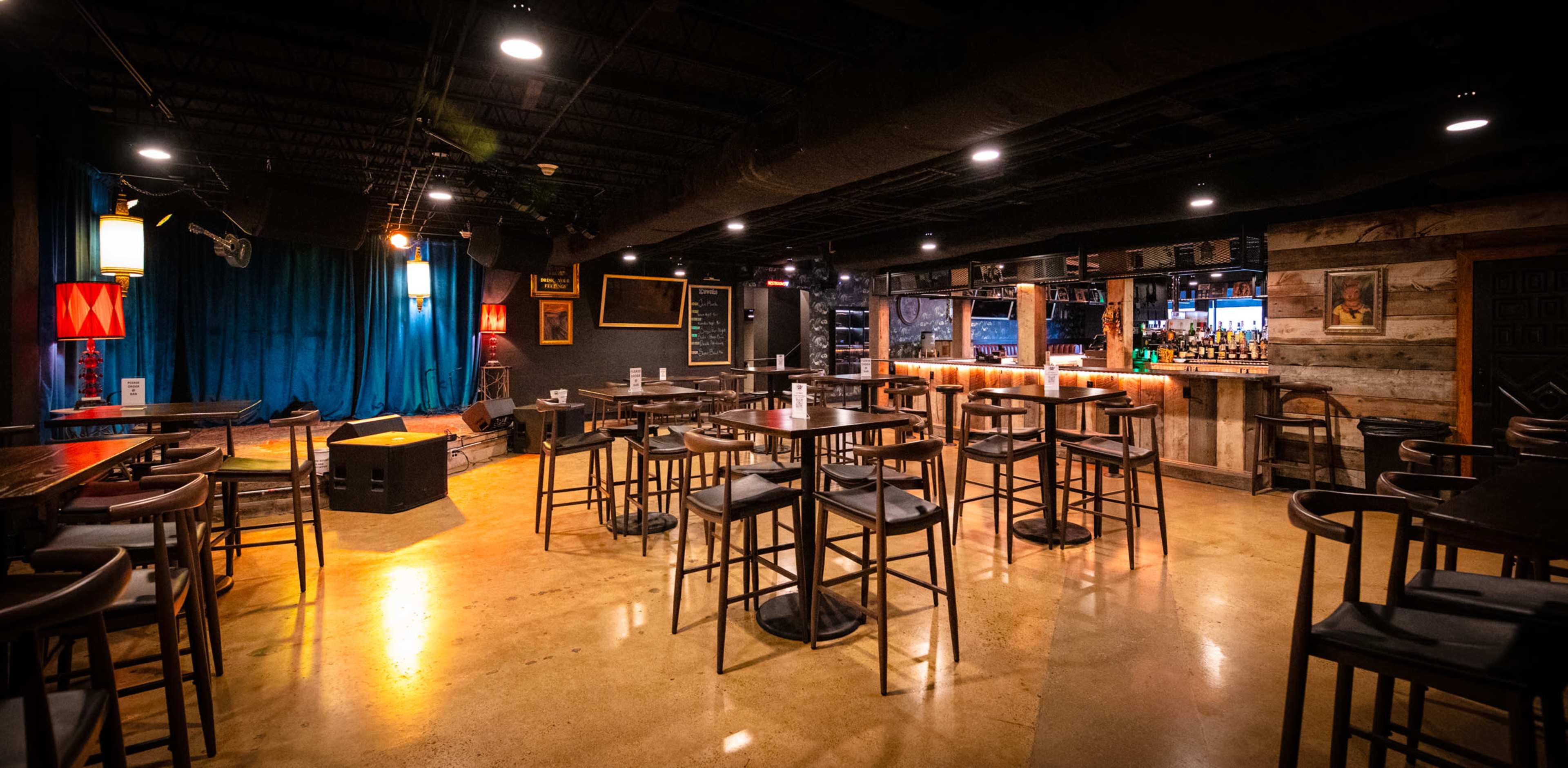 The image shows a cozy bar with wooden tables and stools, vibrant blue curtains, and a well-stocked back bar illuminated by warm light.