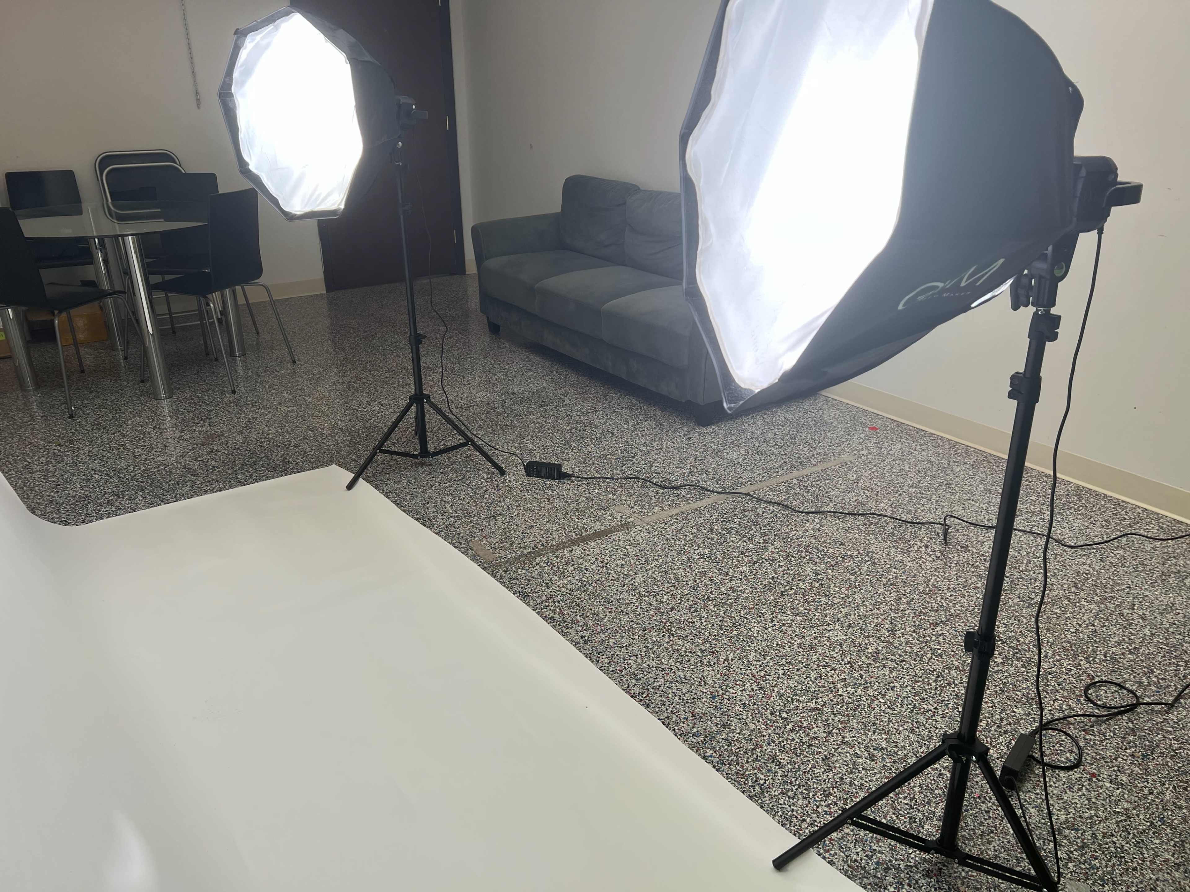 A photography studio setup with two softbox lights on stands, a white backdrop on the floor, and furniture in the background.