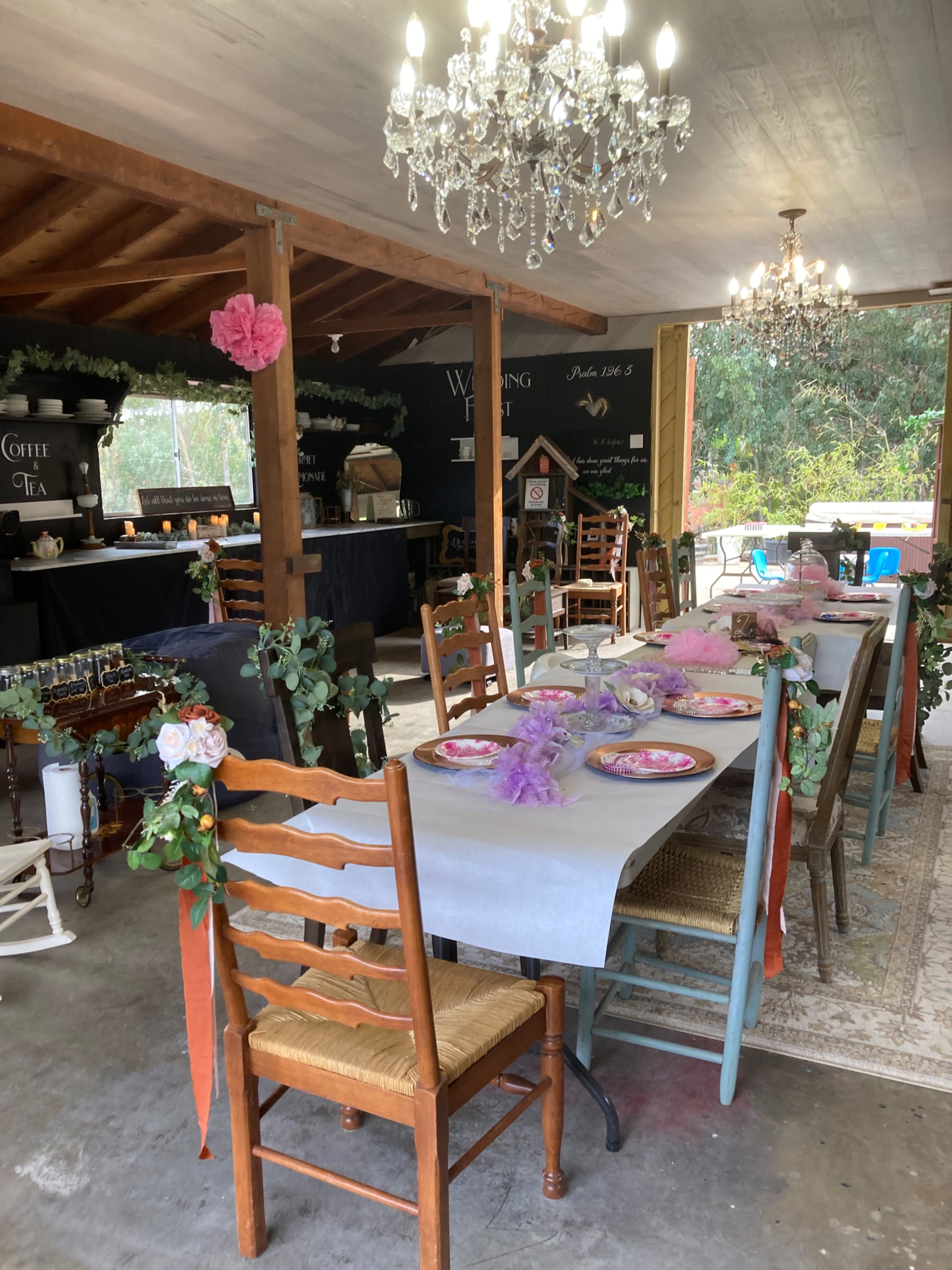A decorated indoor space features tables set with plates and floral arrangements, surrounded by wooden chairs and chandeliers.
