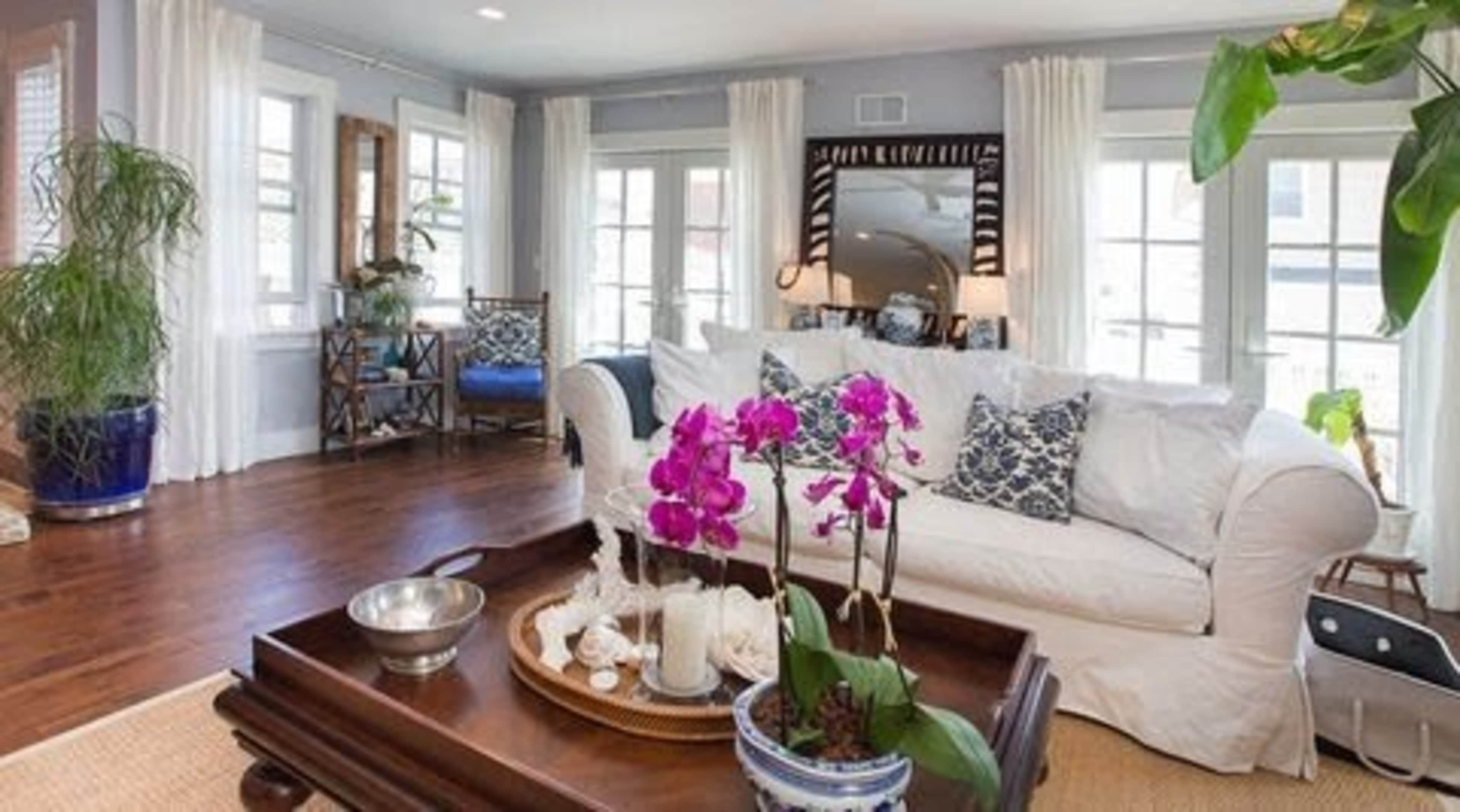 A cozy living room features a white sofa with colorful throw pillows, a wooden coffee table adorned with an arrangement of orchids, and large windows dressed with light curtains.