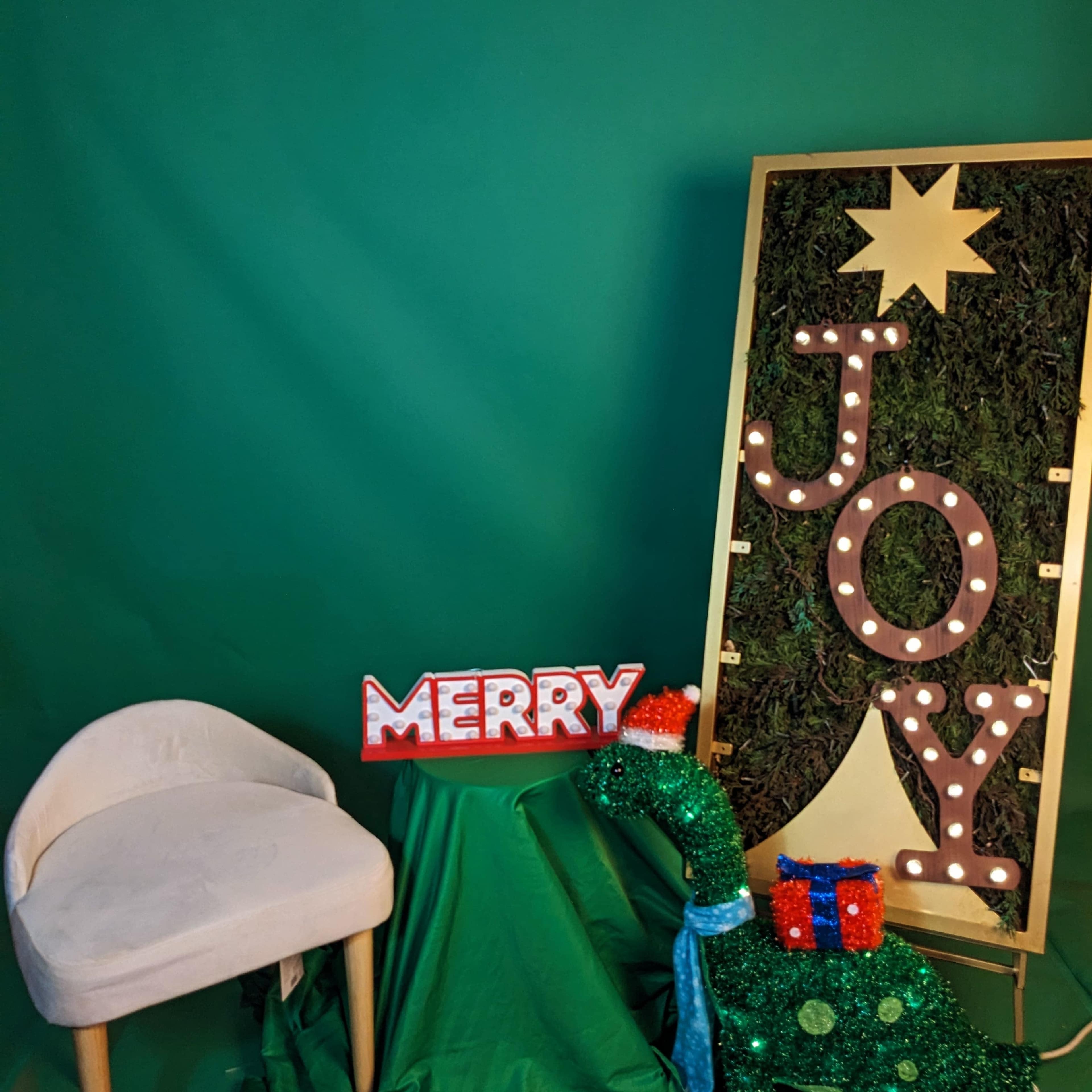 The scene features a green backdrop with a decorative sign reading "JOY," adorned with lights, alongside a chair and a "MERRY" sign.