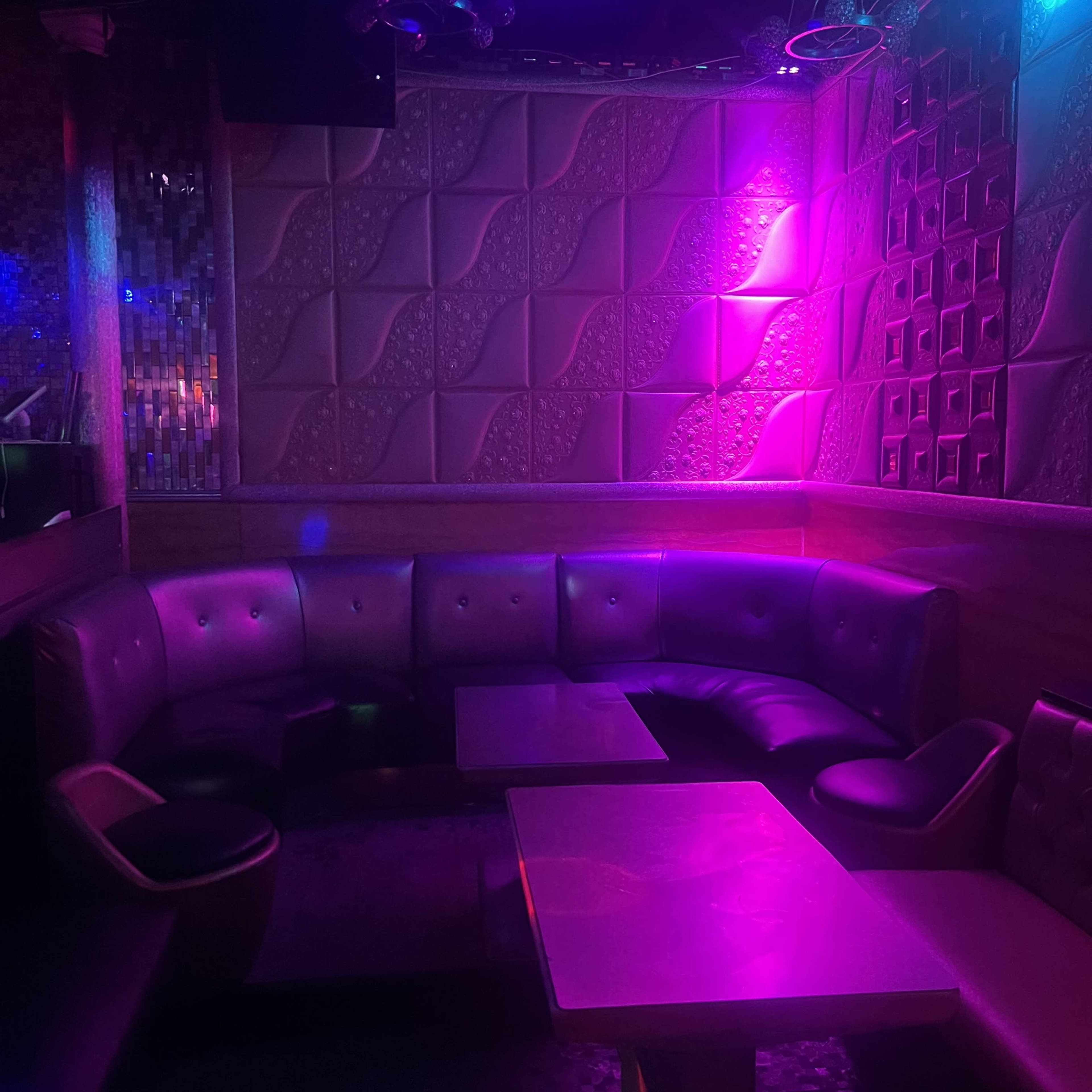 A dimly lit lounge area features curved leather seating and small tables, with colorful lights casting vibrant hues on the textured walls.