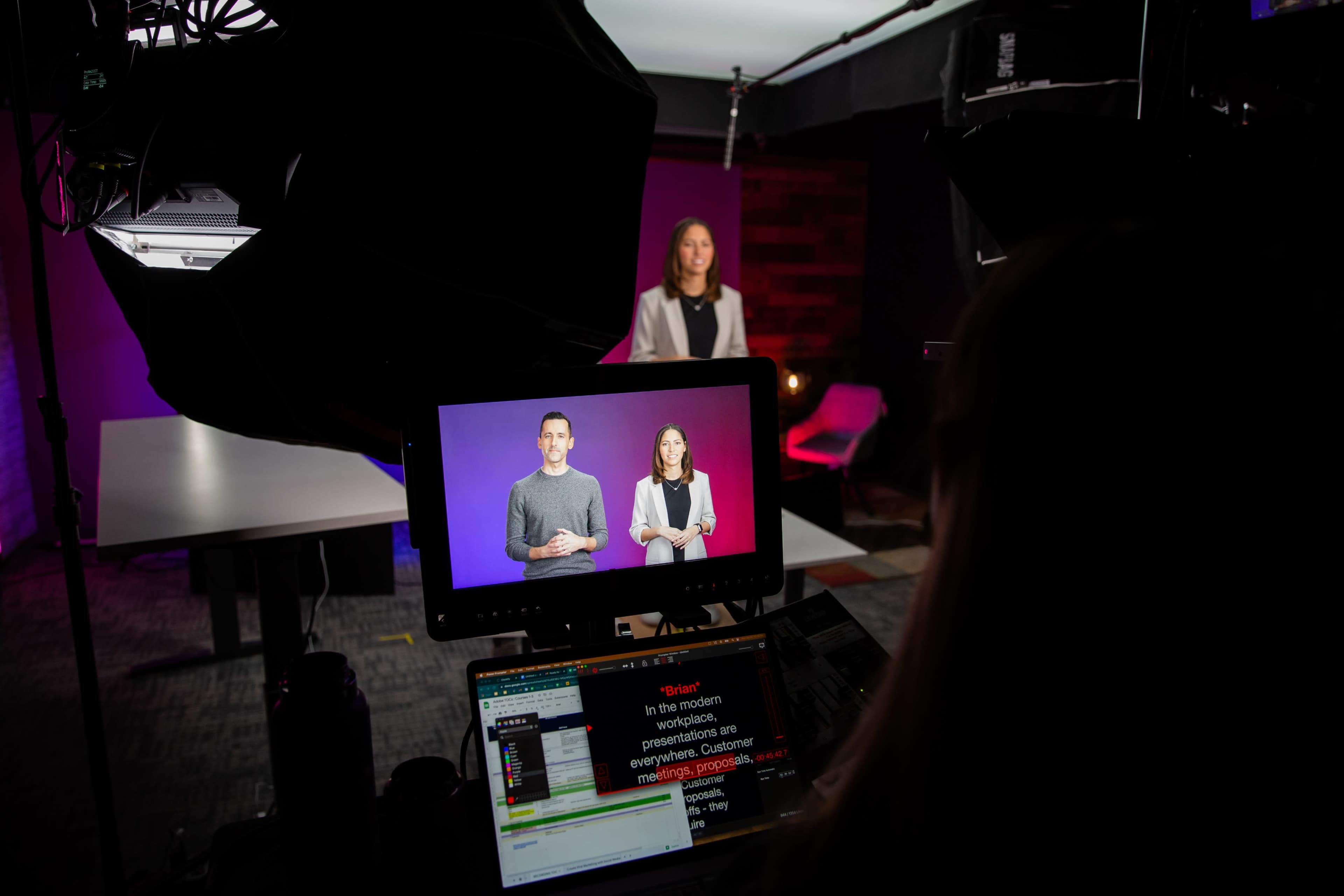A camera setup captures two presenters projected on a screen, while a third person is positioned in front of the camera in a studio environment.