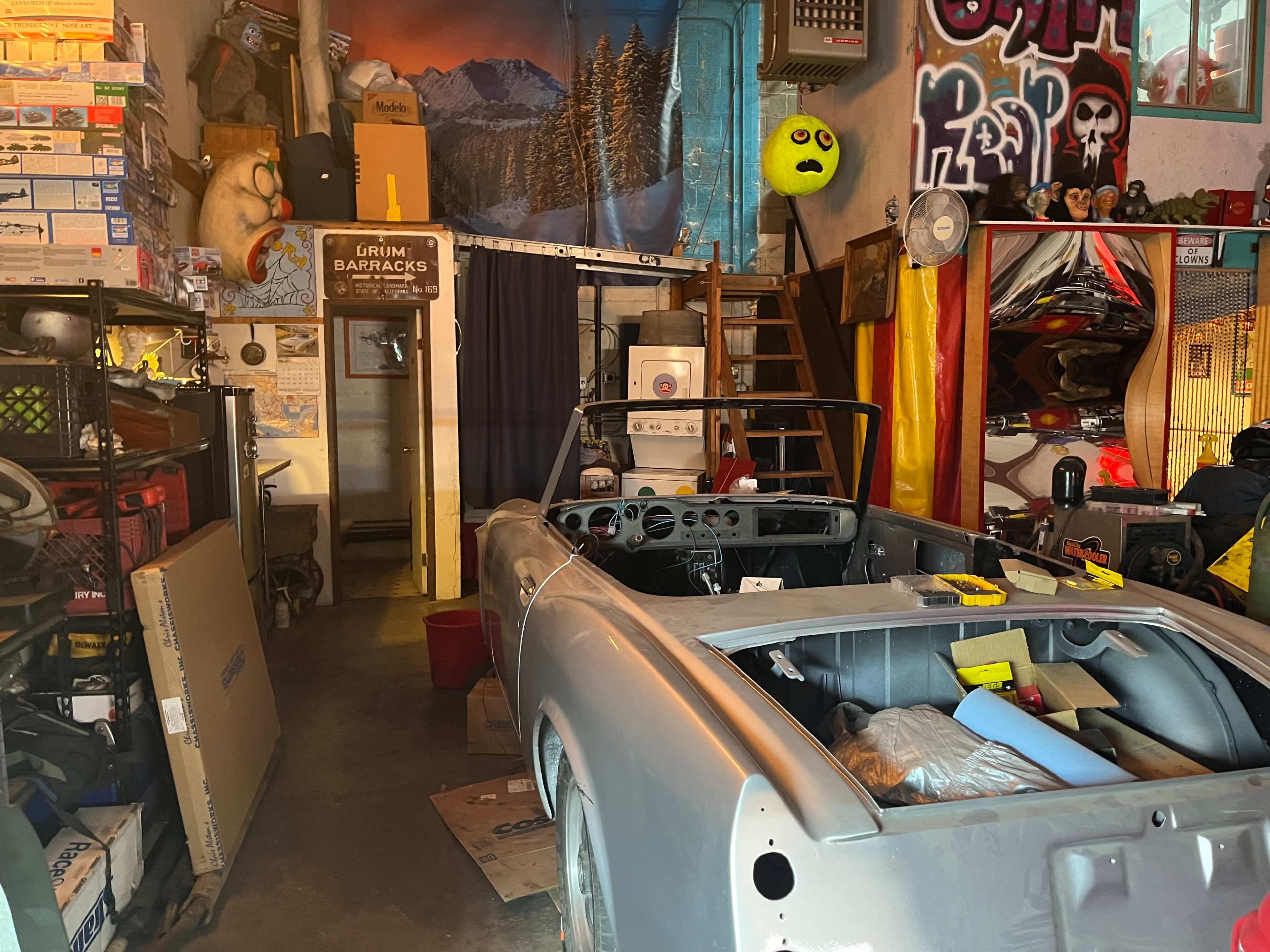 A cluttered garage with a partially assembled gray convertible car in the foreground, surrounded by various tools, boxes, and colorful decorations on the walls.