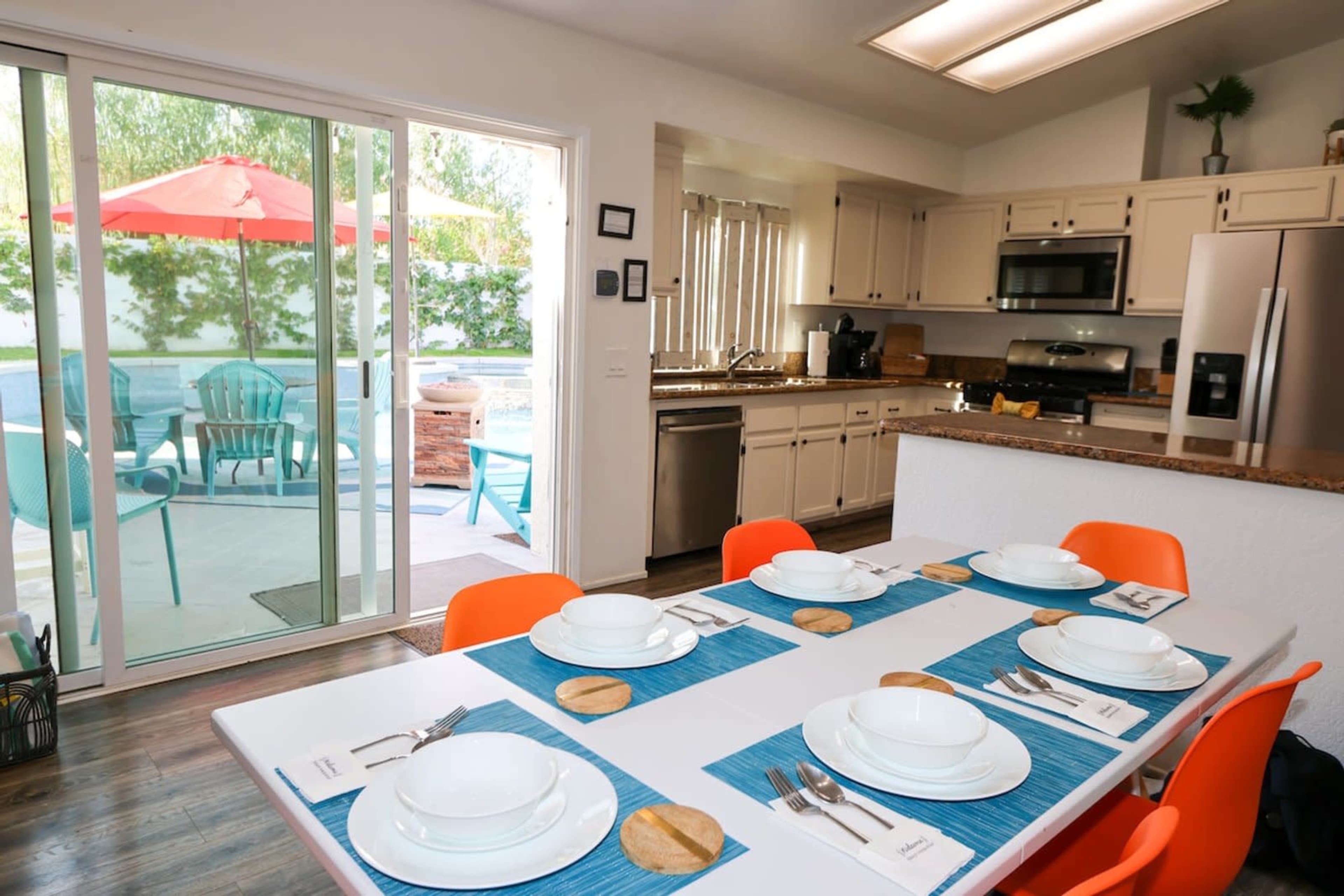 A dining table is set with plates and utensils in a kitchen that opens up to a patio with colorful chairs and an outdoor umbrella.