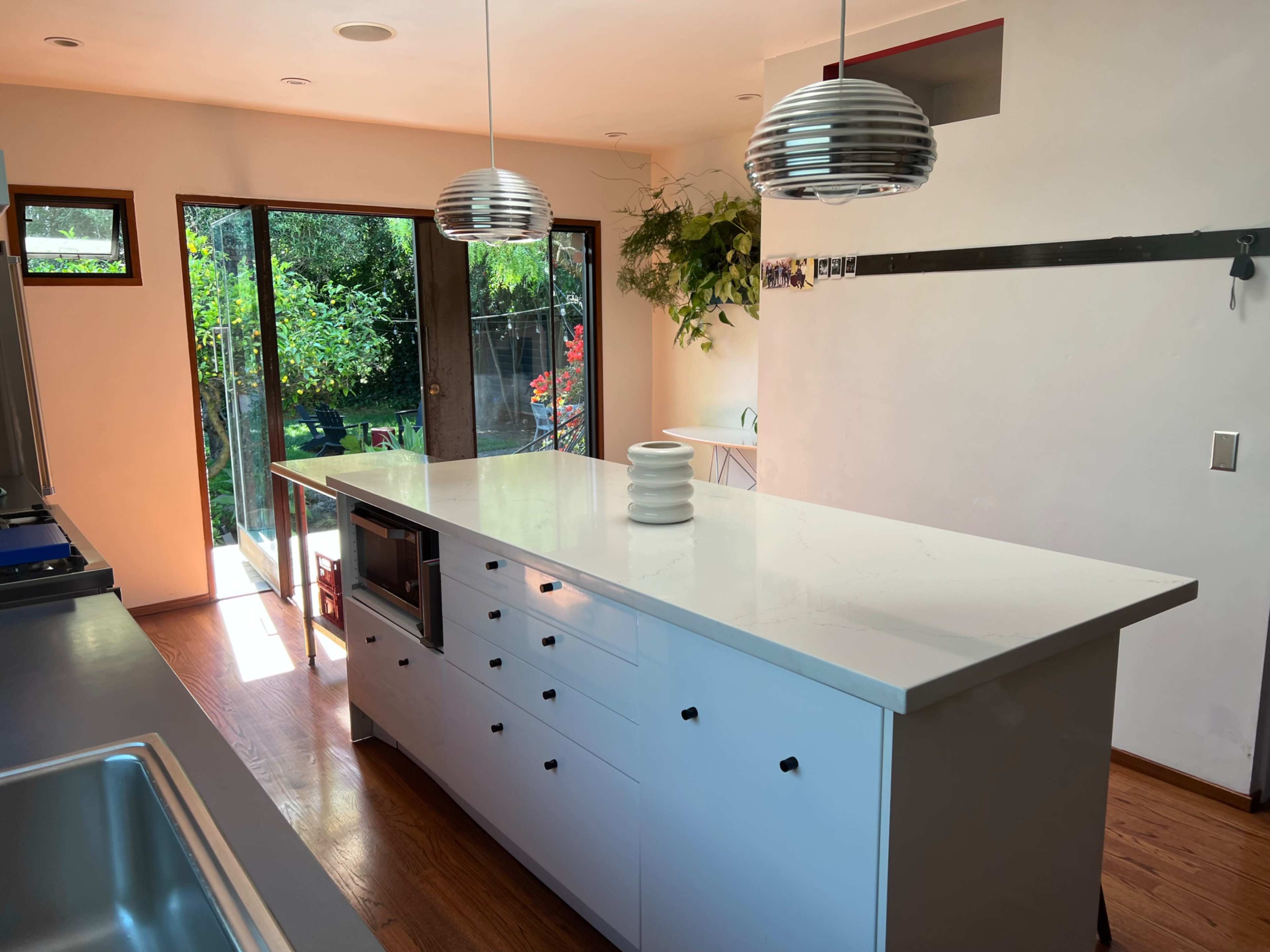 A kitchen features a large, white island with drawers and a minimalist design, overlooking a garden through glass doors.
