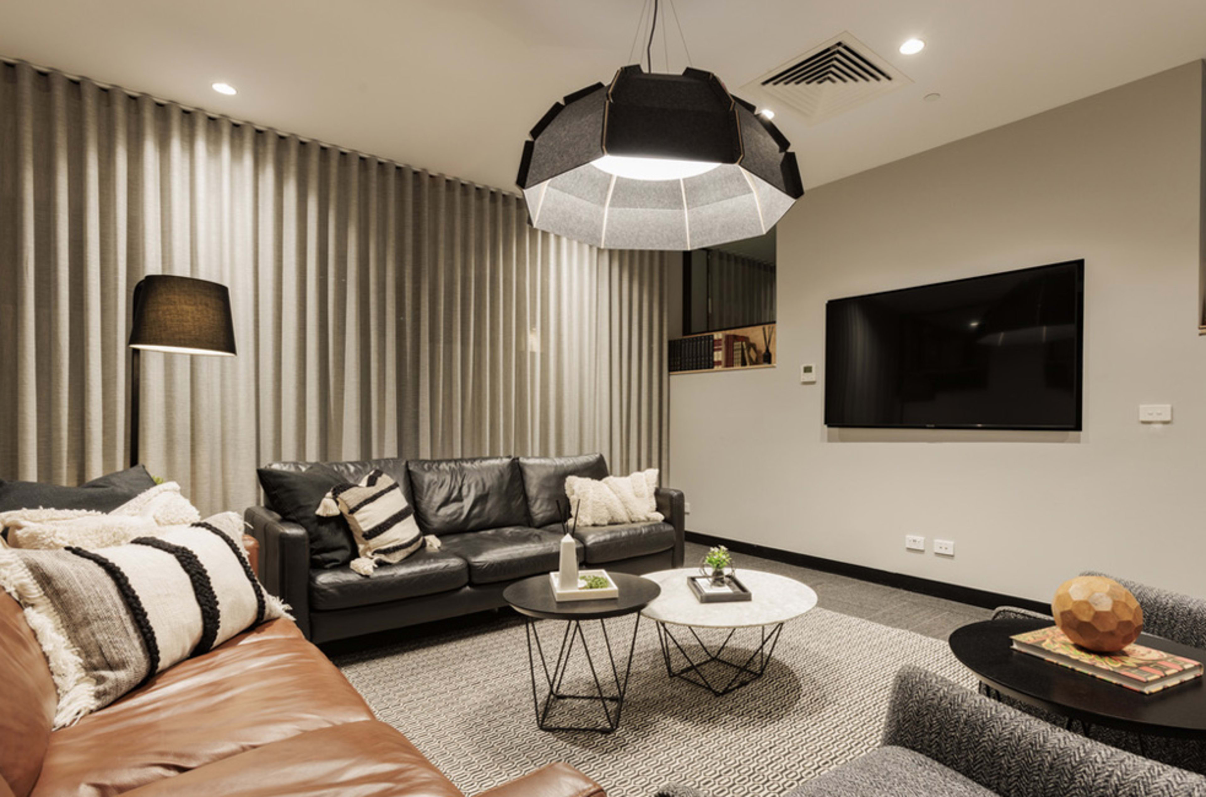 A modern living room featuring a black leather sofa, a brown leather couch, a large pendant light, and two coffee tables placed on a textured rug, with a television mounted on the wall.