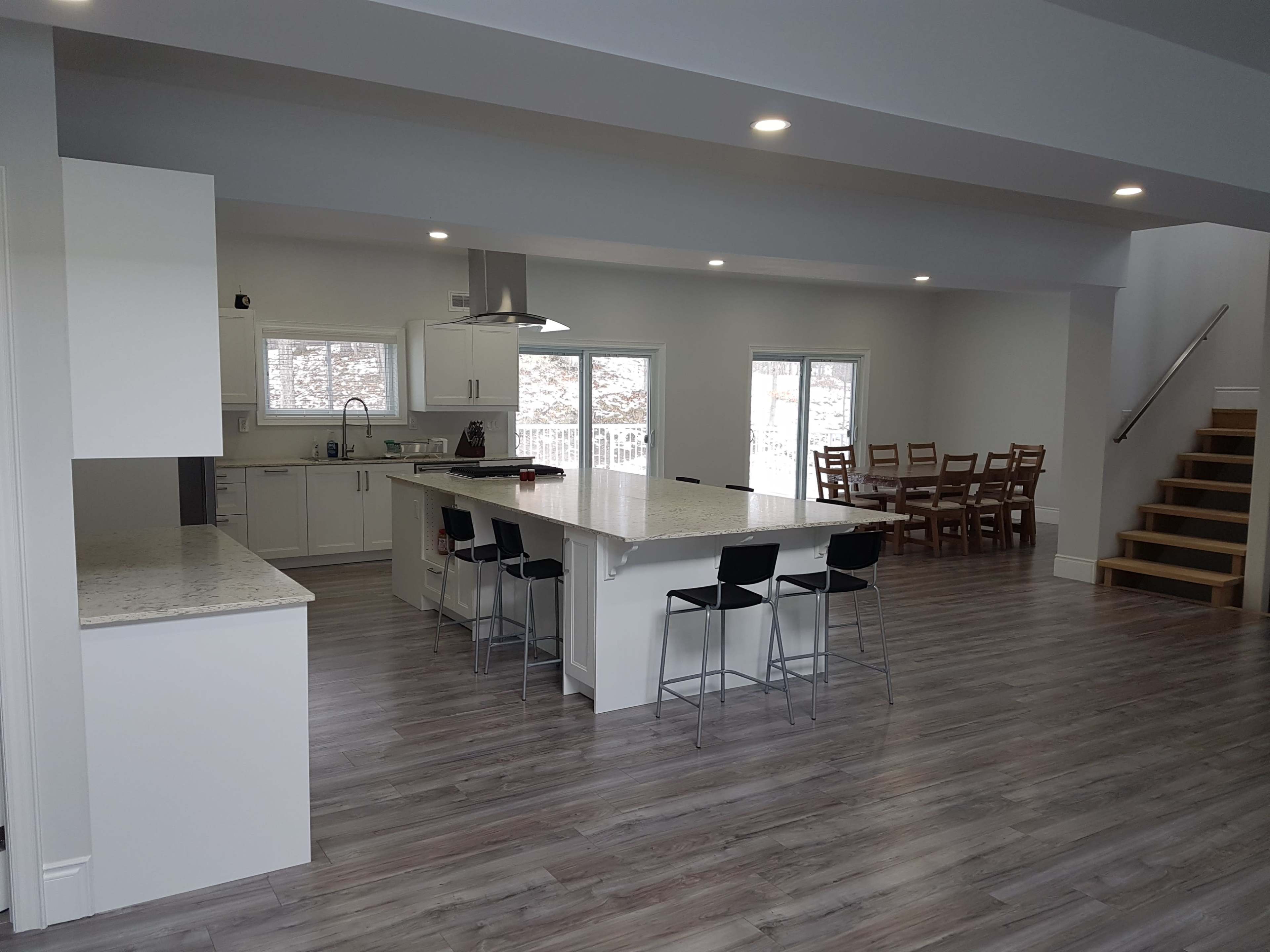 A modern, open-concept kitchen and dining area featuring a large island with bar stools, a wooden dining table, and large windows allowing natural light.