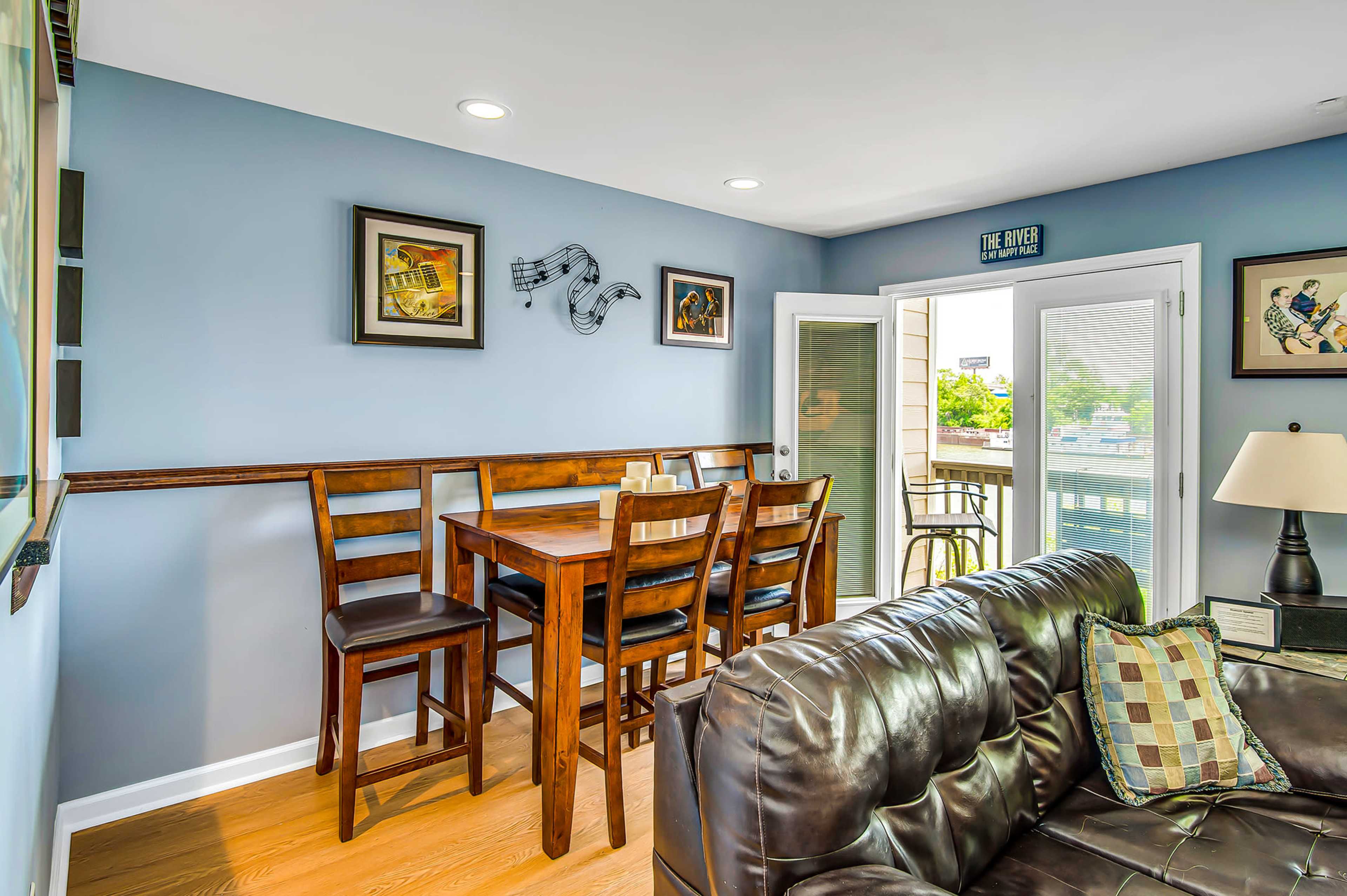 A dining area with a wooden table and chairs is positioned against a blue wall, featuring framed artwork and a door leading to an outdoor area.