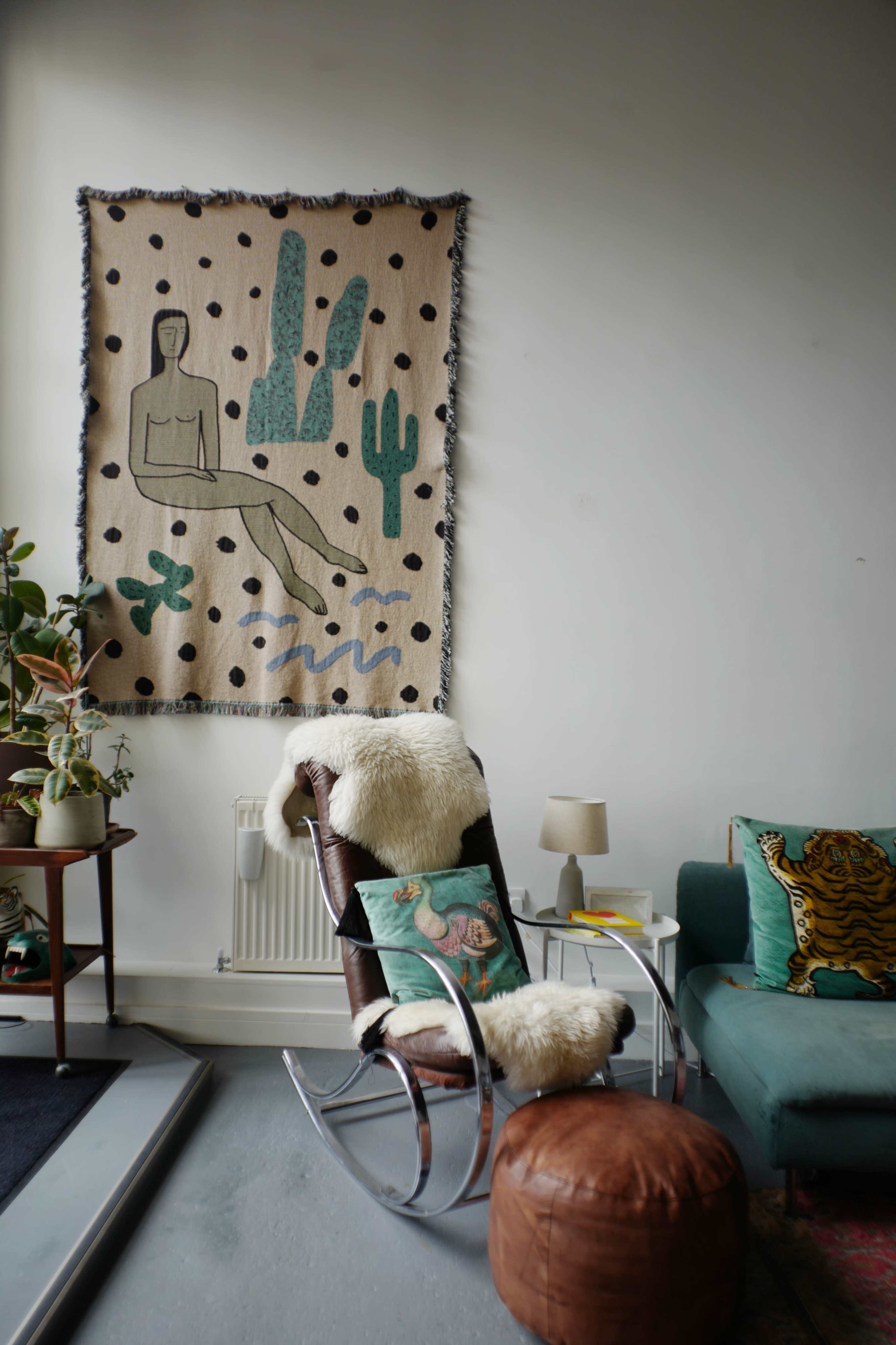 The image shows a cozy interior with a rocking chair, a small table with a lamp, and a wall adorned with a decorative tapestry featuring a figure and cacti.