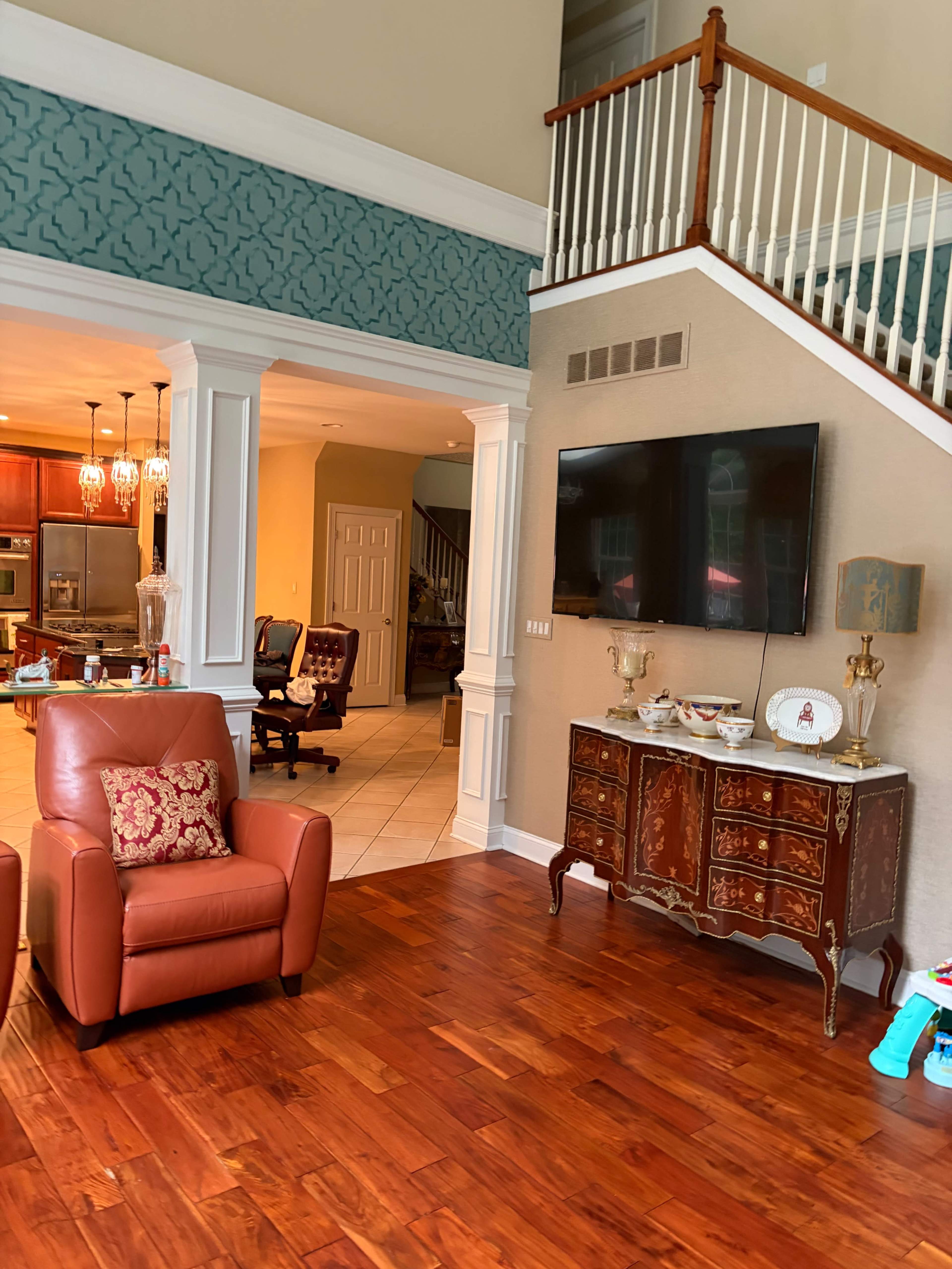 The image shows a spacious living area featuring a pair of brown armchairs, a television mounted on the wall, and a staircase leading to an upper level, with a glimpse of a kitchen and dining area in the background.