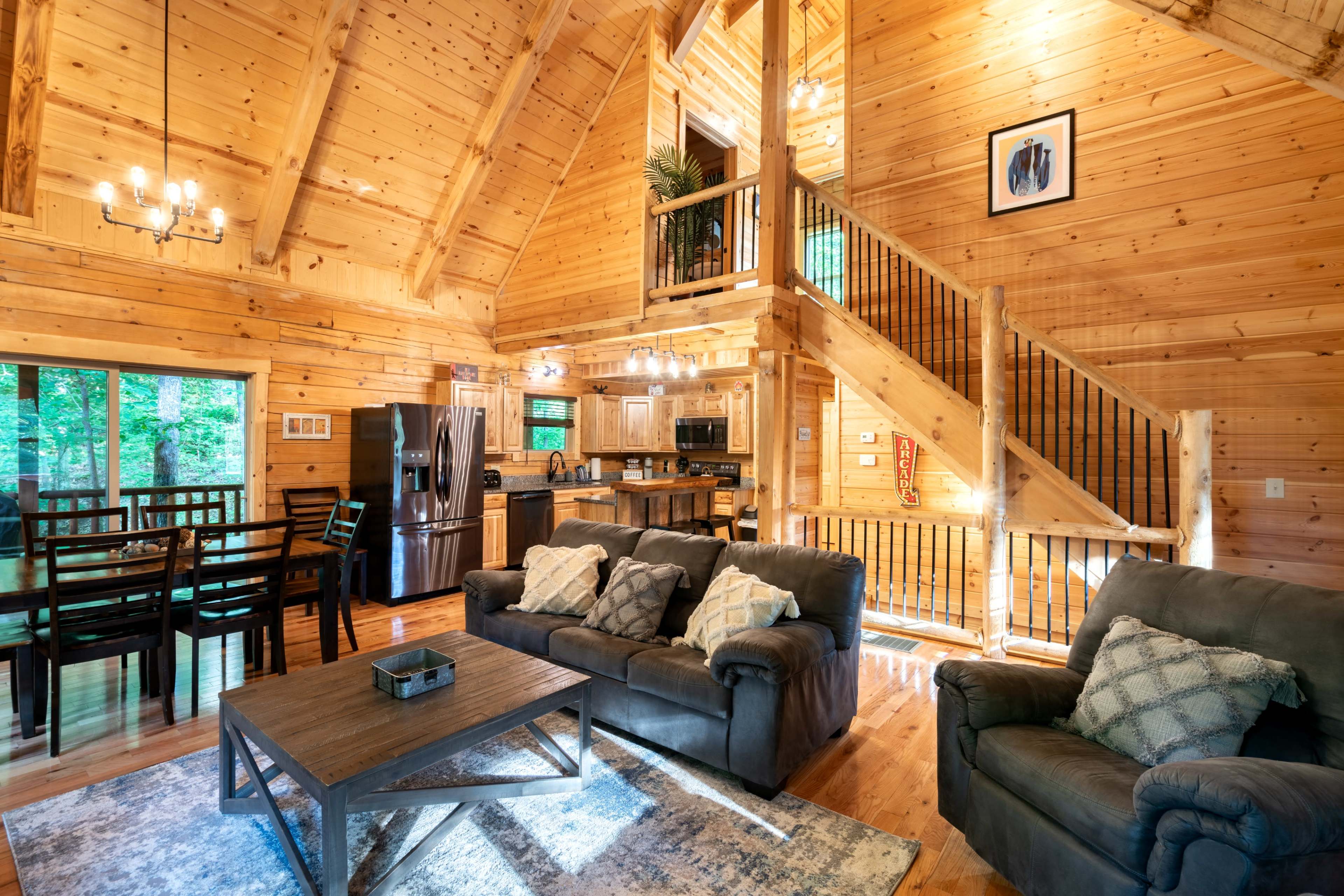 The interior of a wooden cabin features a living area with a couch, coffee table, and dining space, alongside a kitchen and staircase leading to a lofted area.