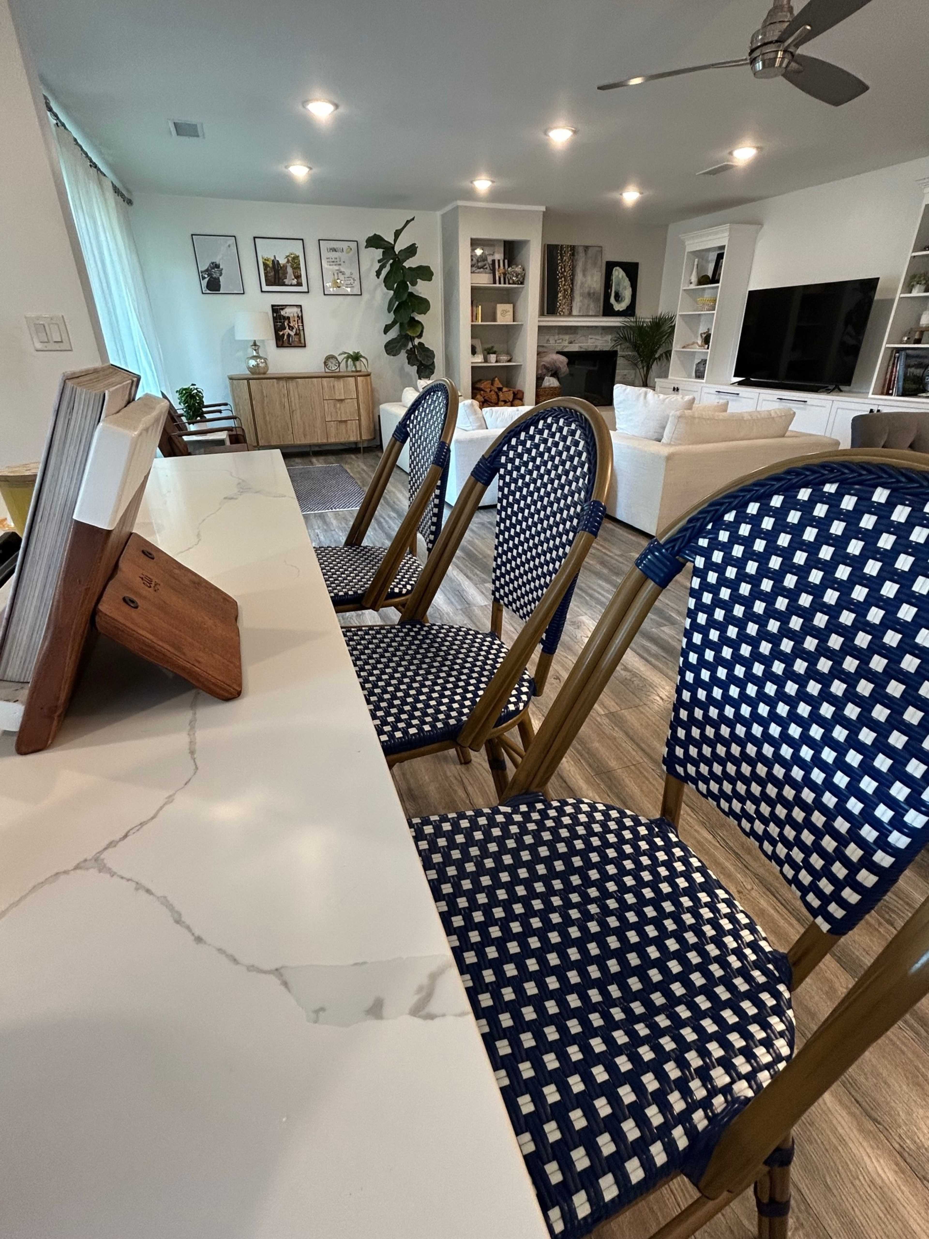 A row of three blue-and-white patterned chairs is positioned at a kitchen counter, facing a modern living room with a sofa and wall-mounted shelves.