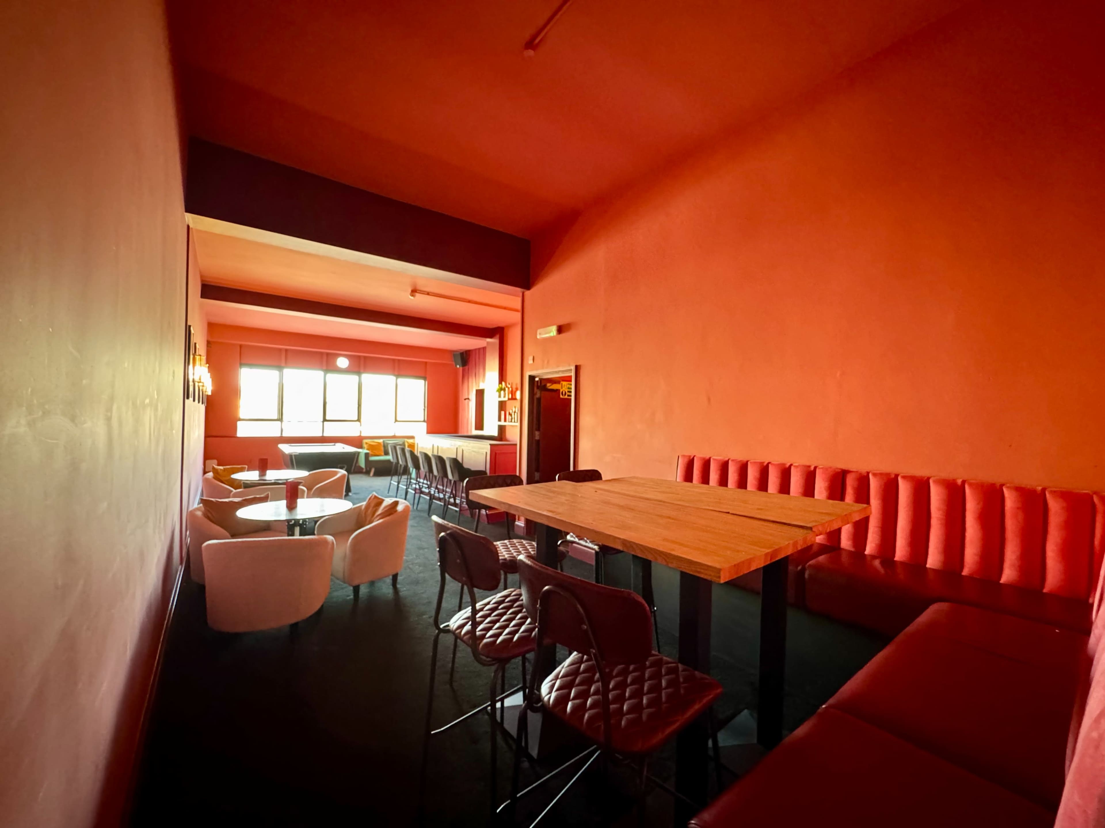 The image shows a brightly colored room with red walls, featuring a mix of high tables and seating areas that create a lounge atmosphere.