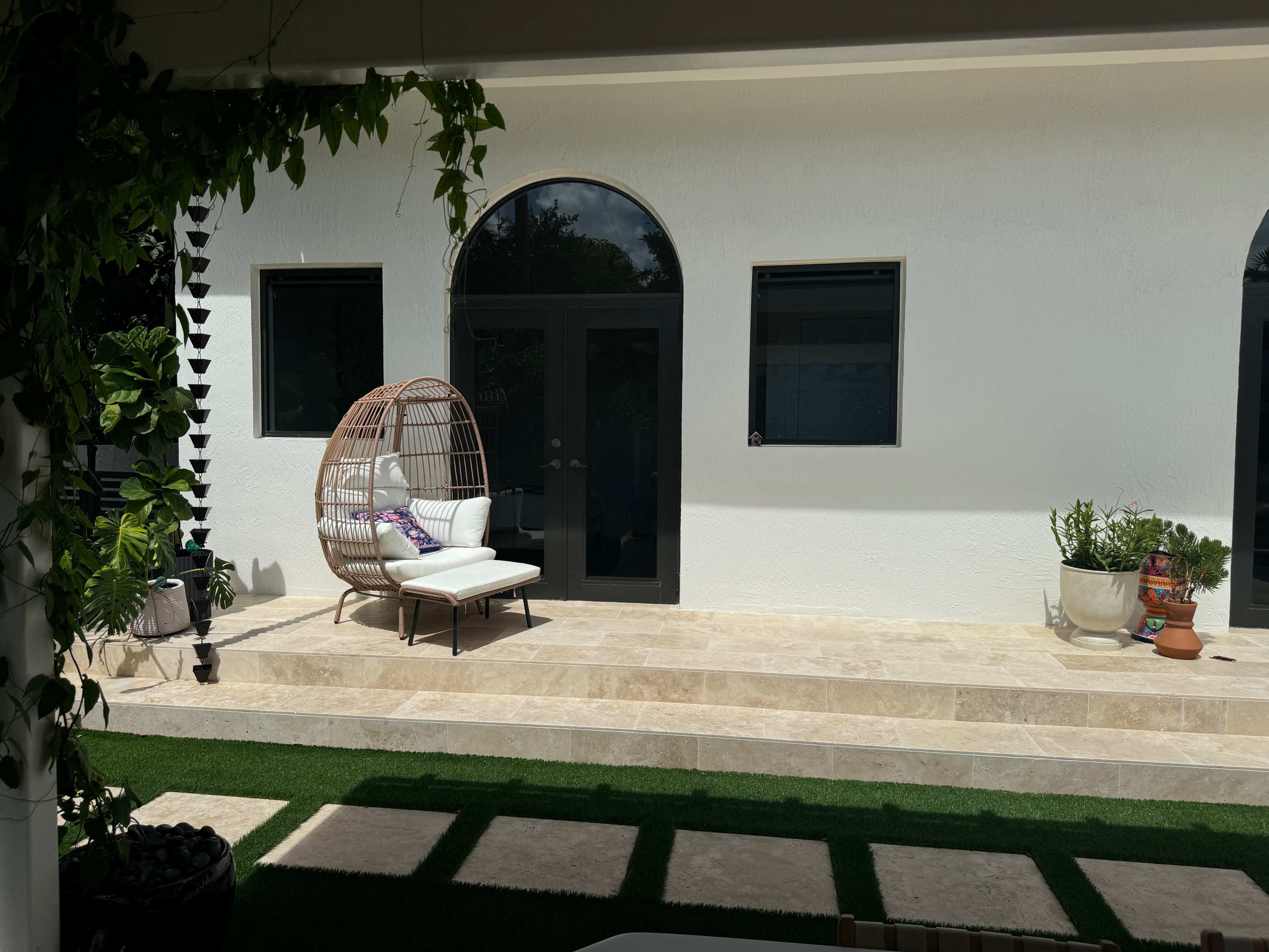 A patio area features a curved-back wicker chair with cushions, positioned near a white wall and surrounded by greenery and potted plants.
