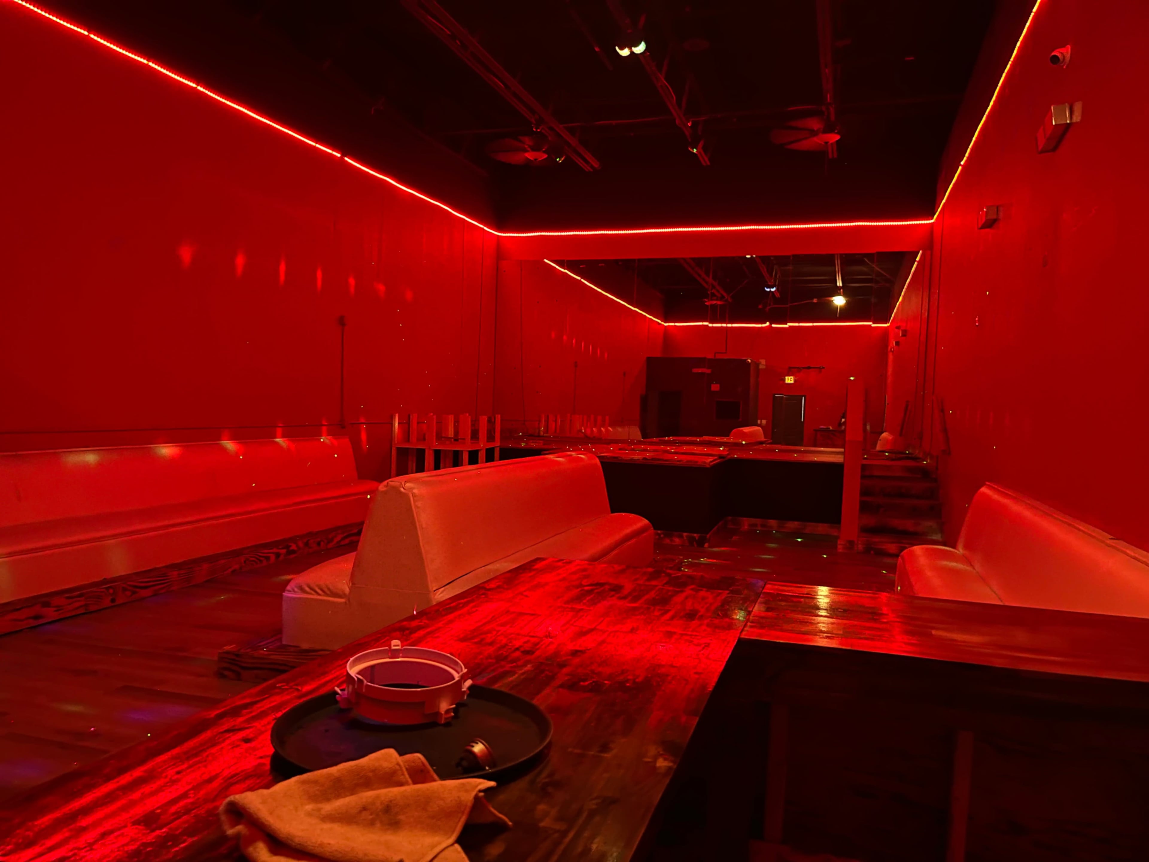The image shows a dimly lit room with red walls, benches, and a wooden table, illuminated by red neon lights.