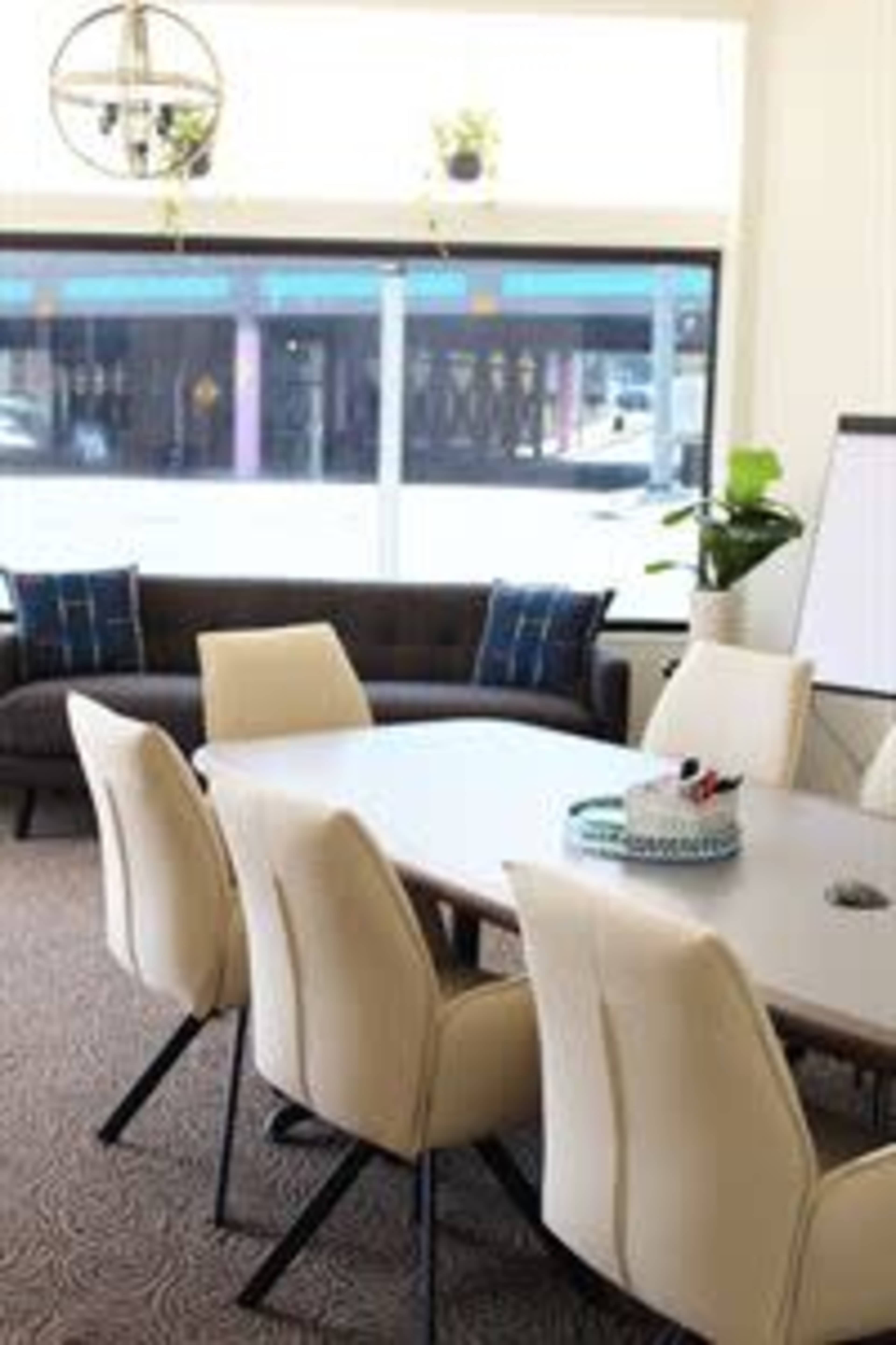 A modern conference room features a large table surrounded by cream-colored chairs, with a couch, plants, and a whiteboard visible in the background.