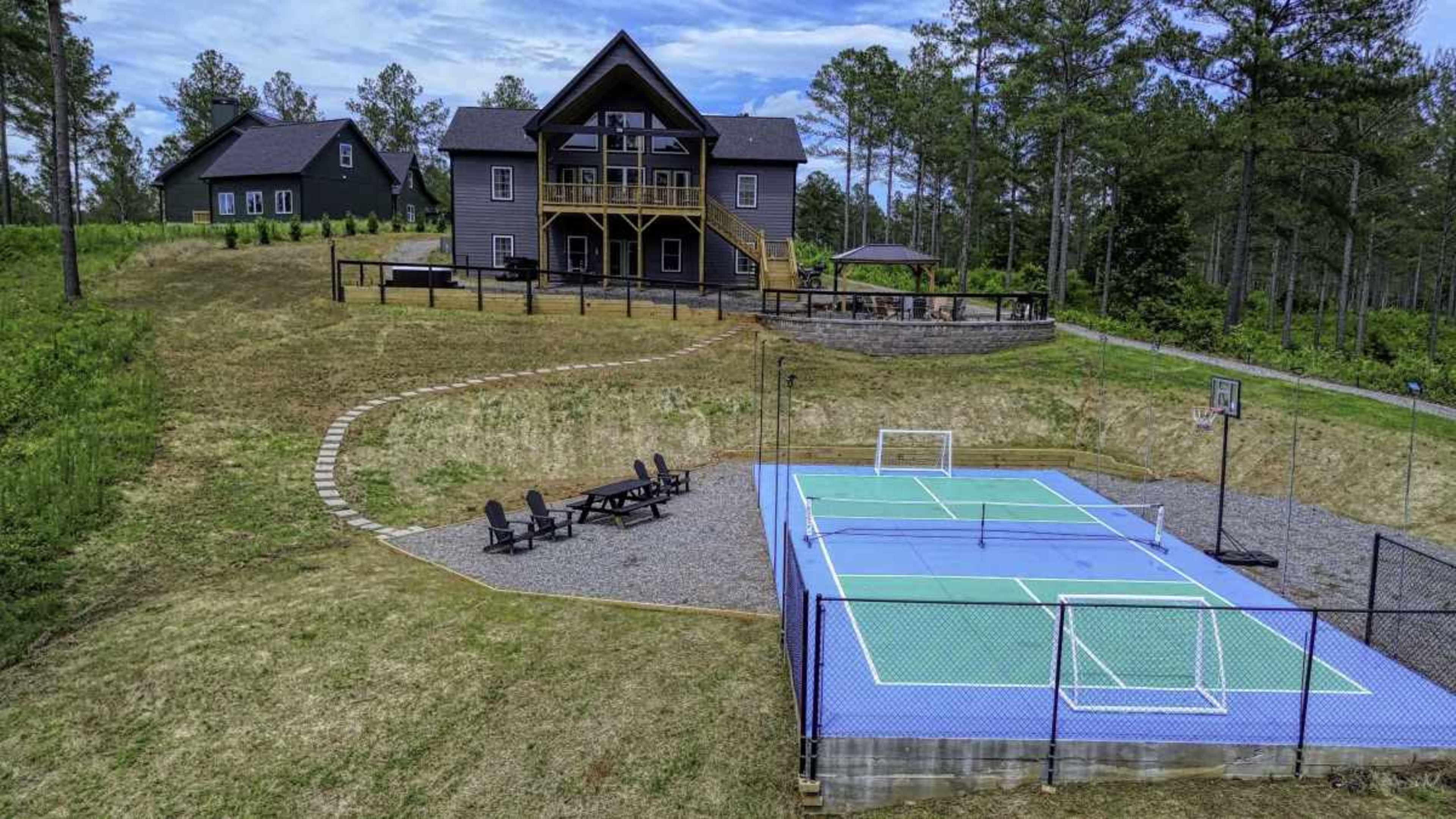 The image shows a large house in the background with a spacious deck, adjacent to a gravel area featuring a multi-sport court marked for tennis and basketball.