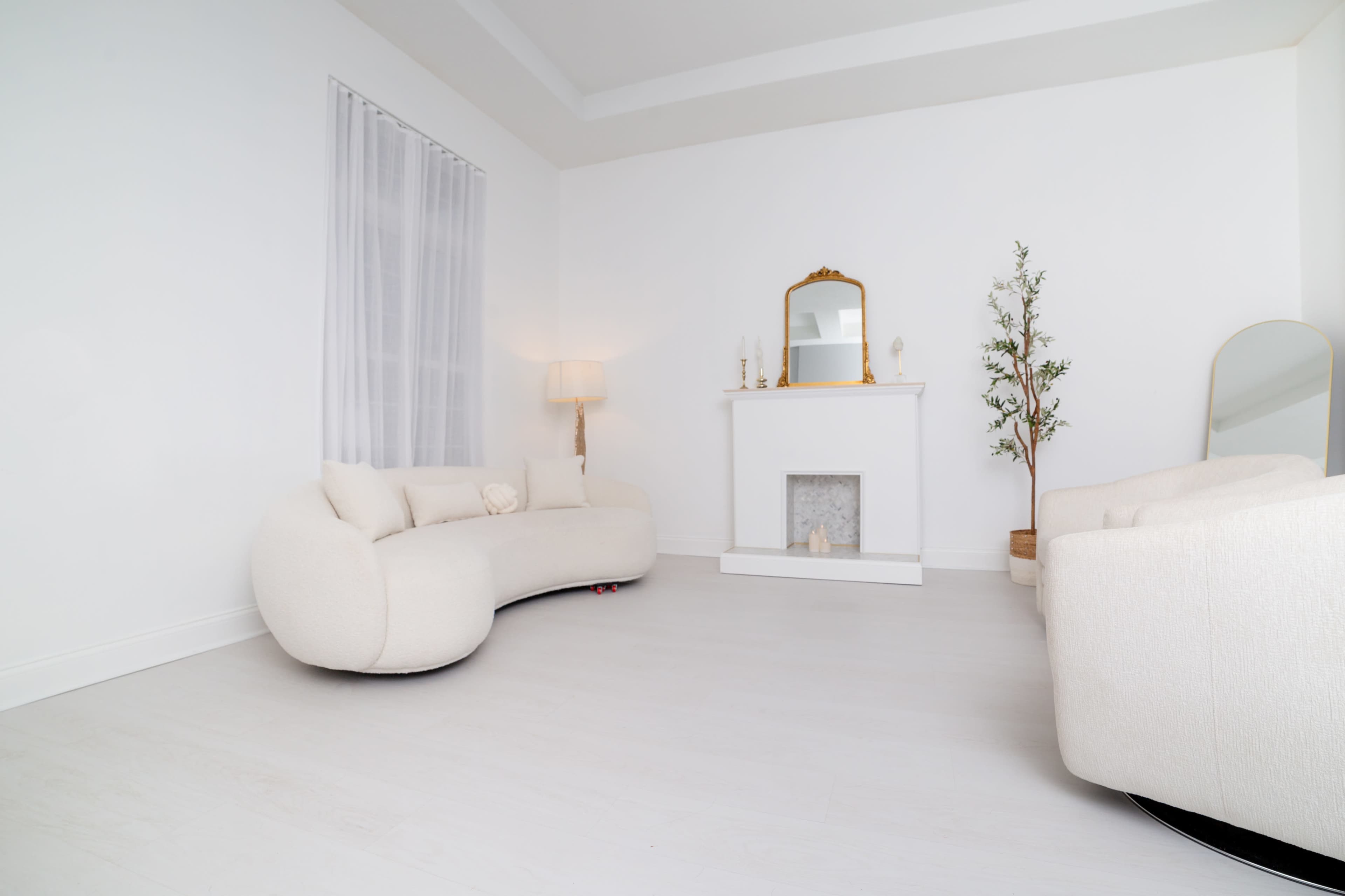 The image shows a minimalist, white-walled living room featuring a curved cream sofa, two armchairs, a lamp, a fireplace with a mirror above it, and a decorative plant.