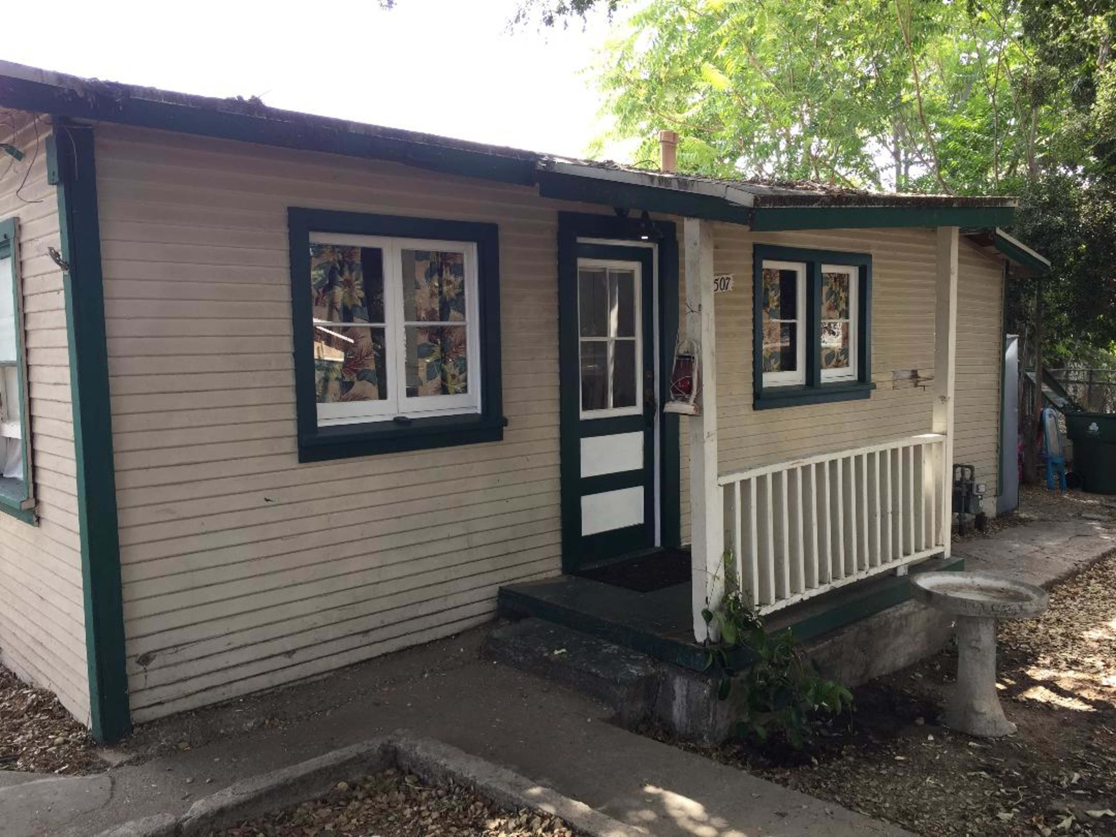 A small, one-story house with light-colored siding and green trim features a front porch and a garden area beside it.