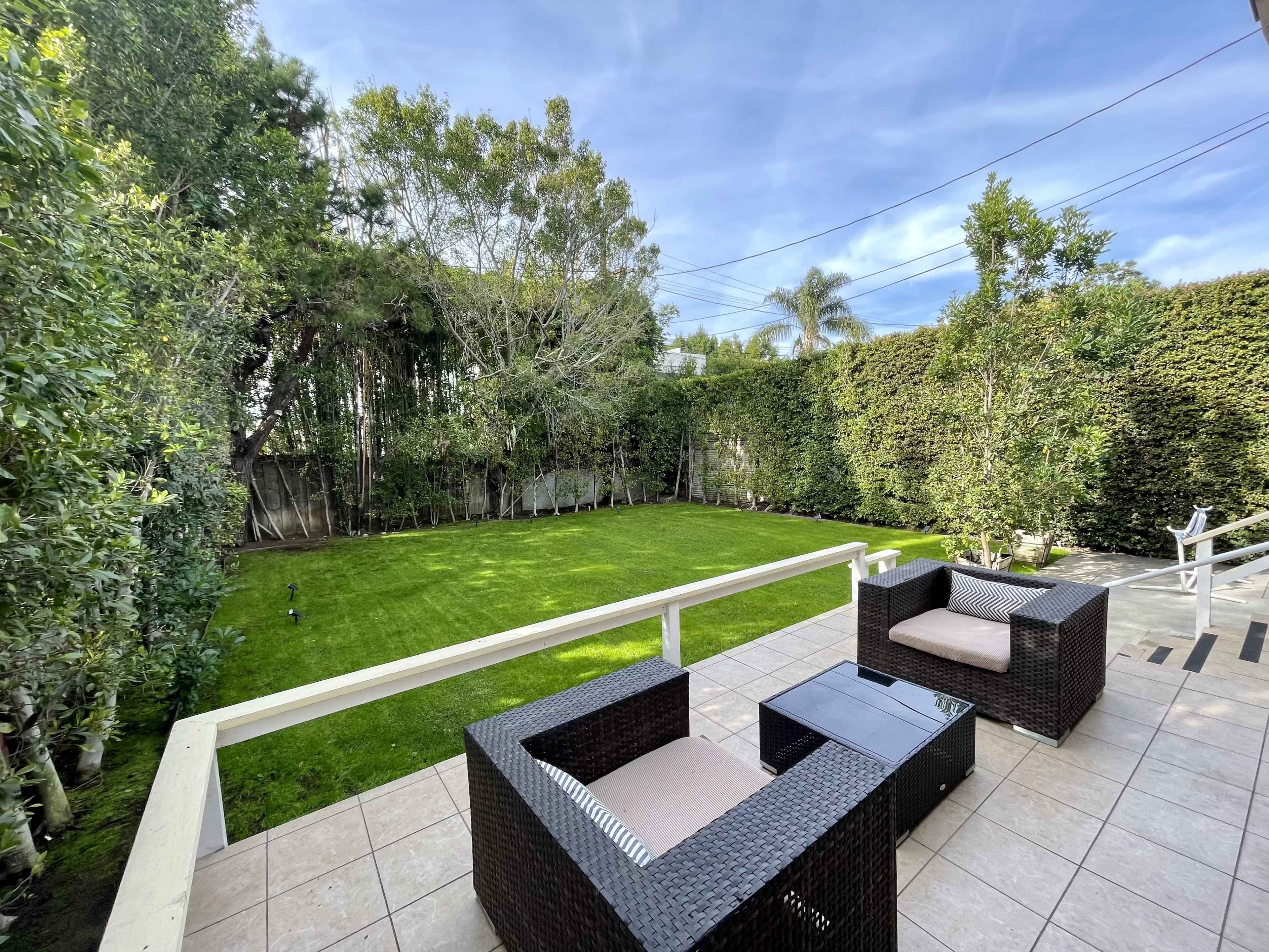 A neatly maintained backyard features a manicured lawn surrounded by tall hedges, with rattan chairs positioned on a tiled patio.