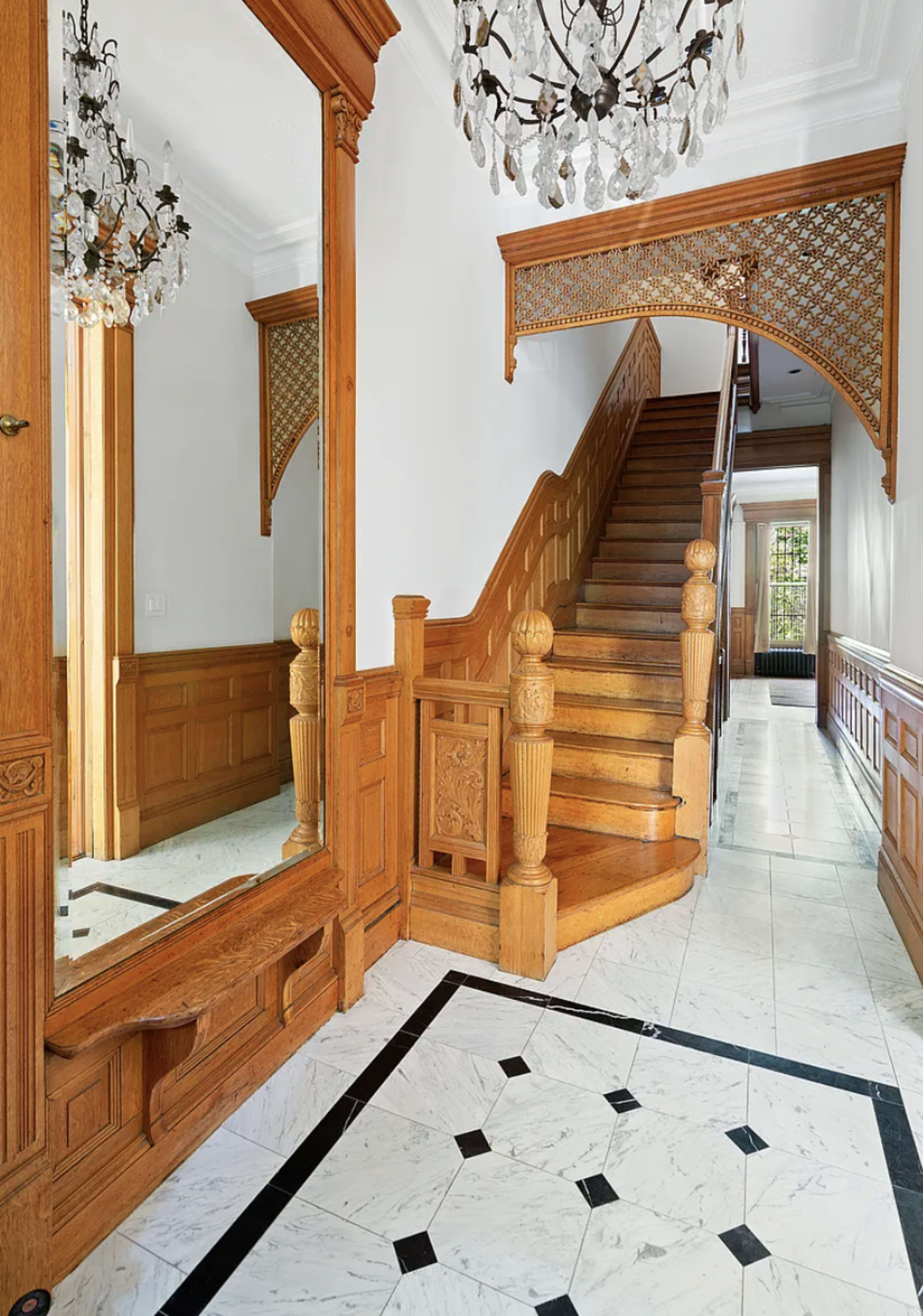 The image features a spacious entryway with a wooden staircase, ornate woodwork, and a large mirror reflecting the surroundings.