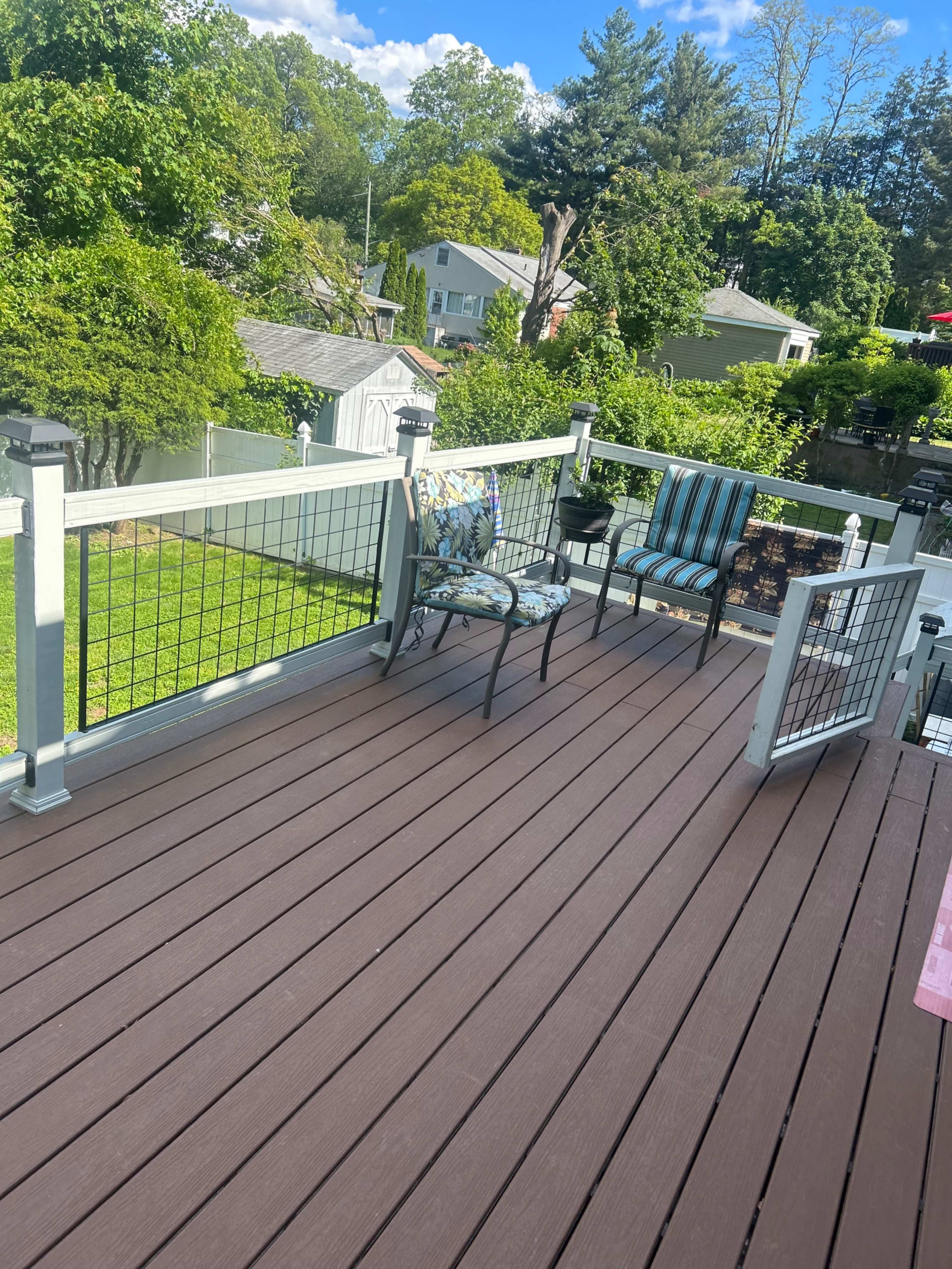 The image shows a wooden deck with two metal chairs and a small table, surrounded by greenery and houses in the background.