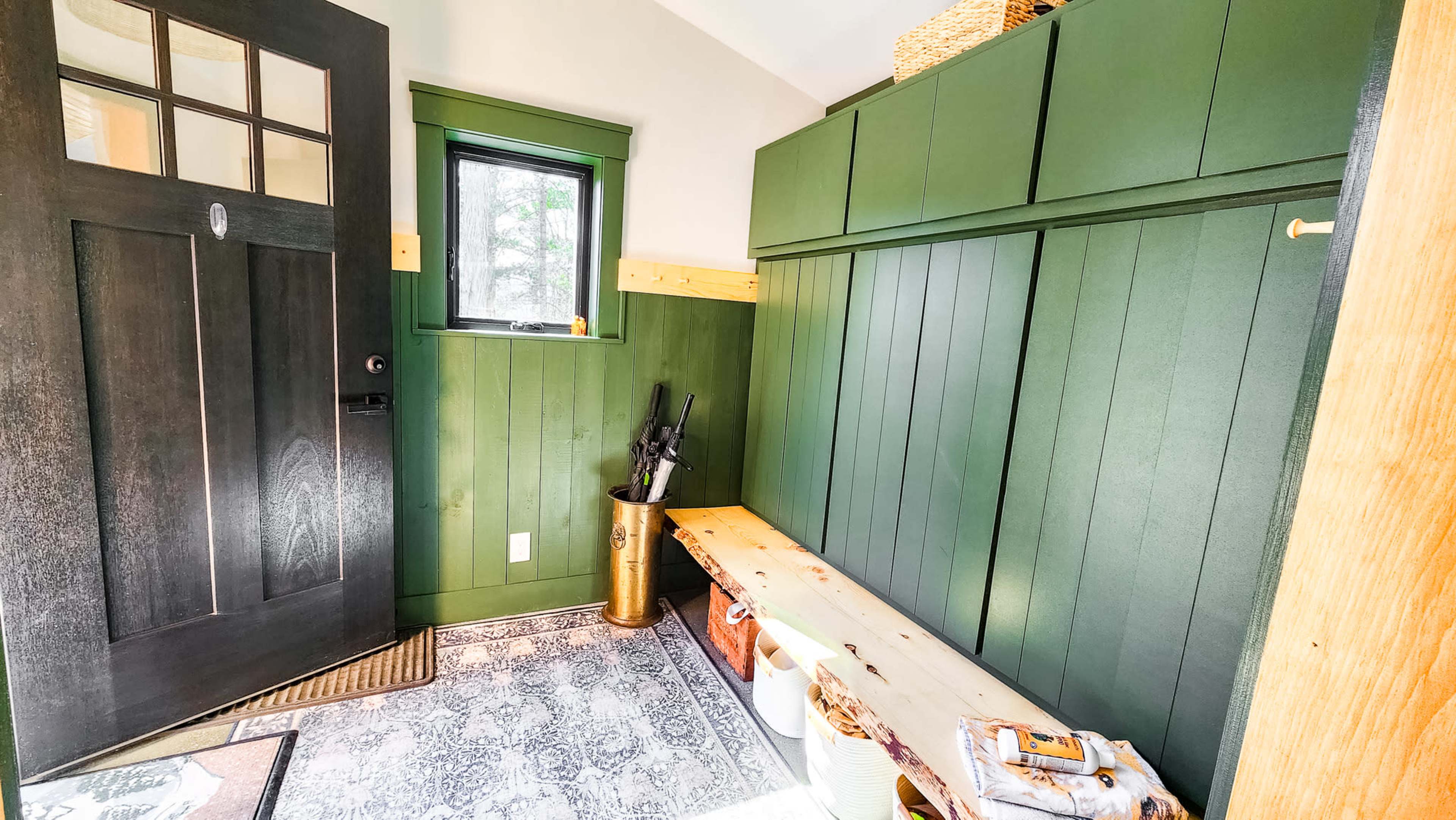 The image shows a mudroom with green paneled walls, a bench, and storage shelves, featuring a door on the left and a window on the right.