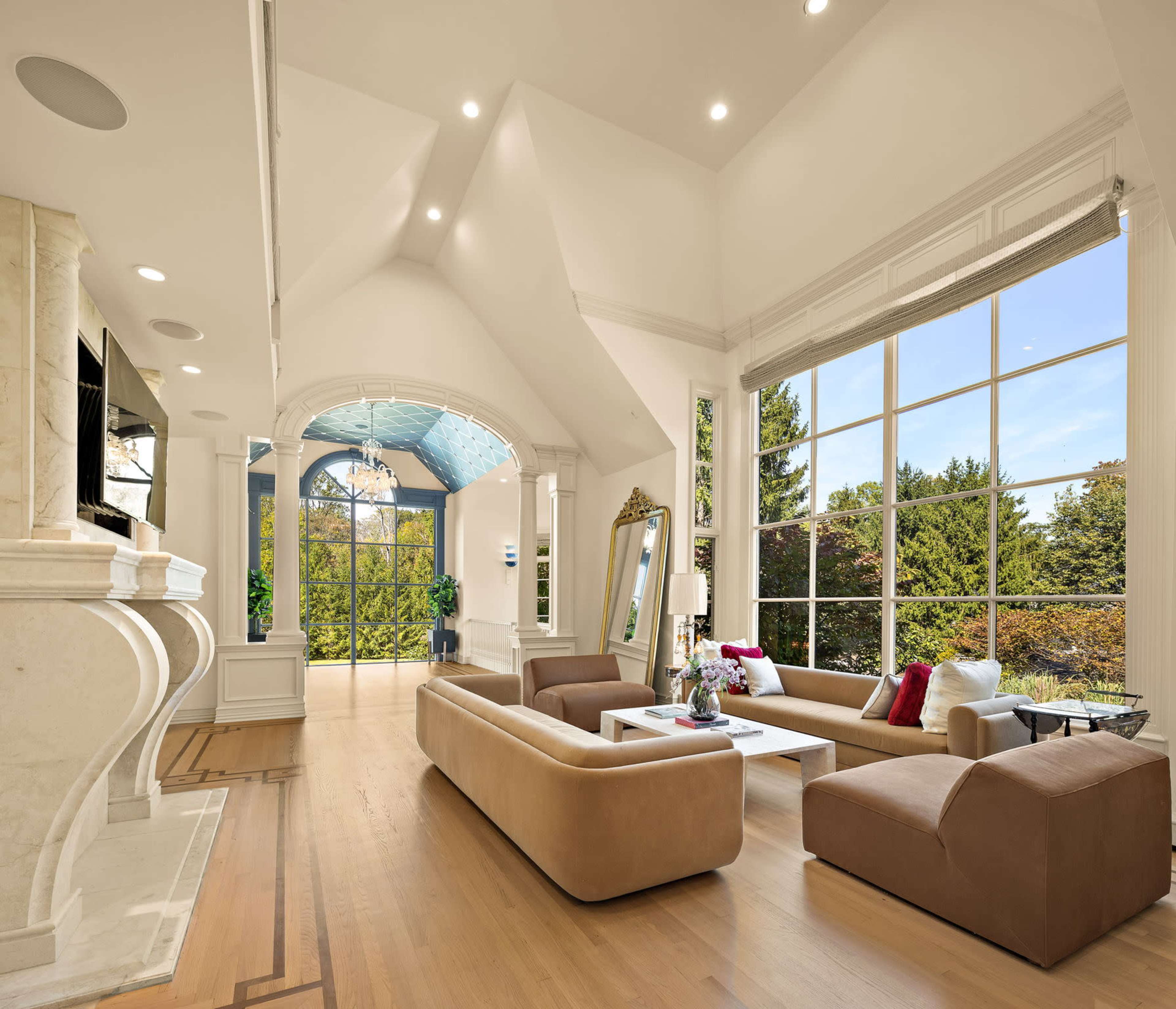 The room features a spacious, modern living area with large windows offering a view of greenery, furnished with two sofas, a coffee table, and a fireplace.